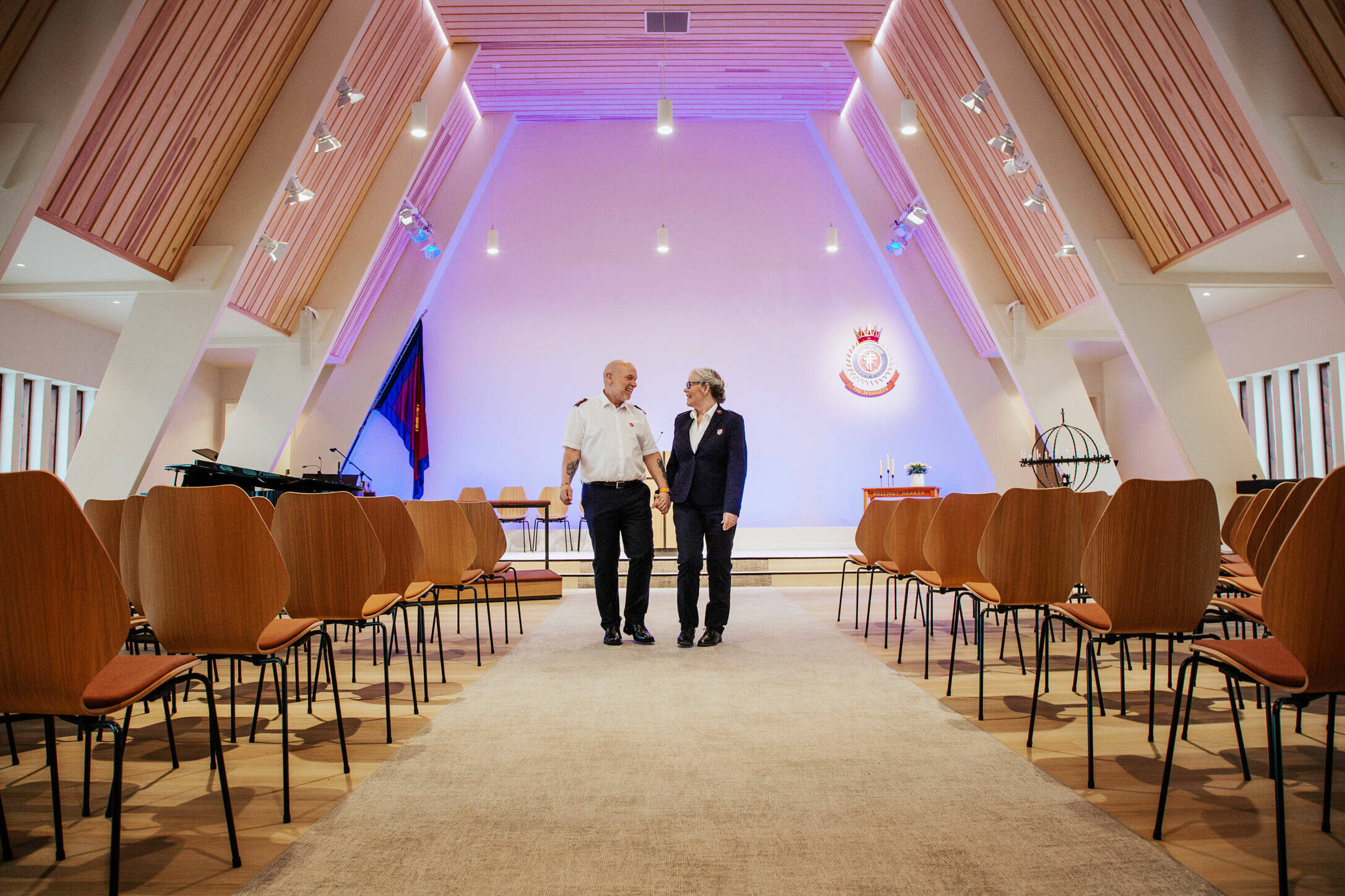 Foto: mann og kvinne i uniformer fra Frelsesarmeen står i et kirkerom med blå belysning i enden av rommet. Veggene er skrå.