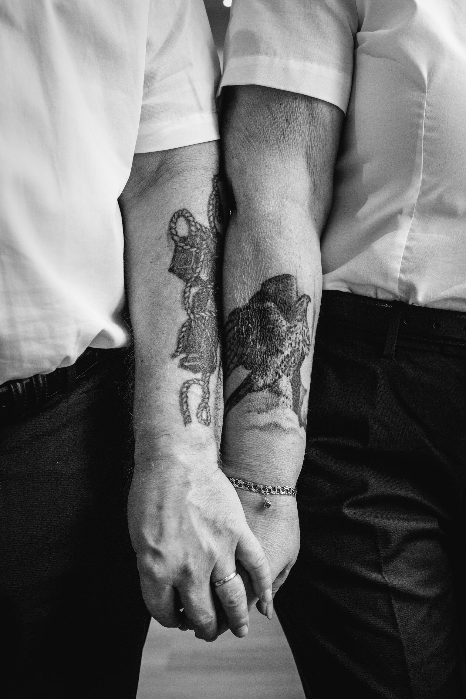 Foto: Kvinnearm og mannsarm med tatoveringer holder hverandre i hendene. Bildet er i sort-hvitt.