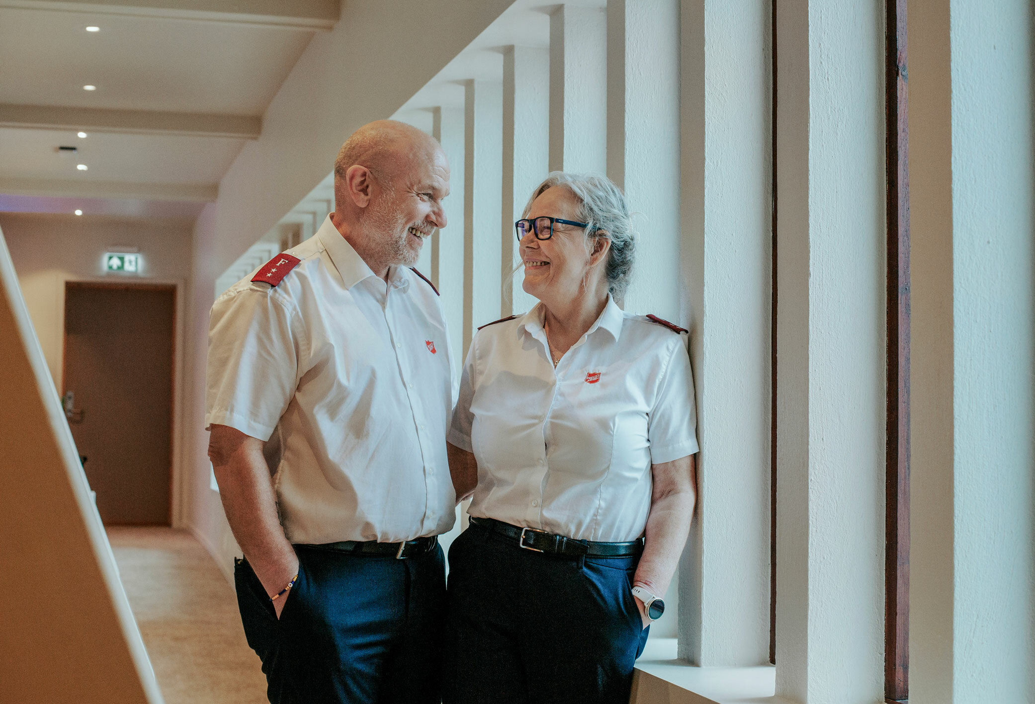 Foto: Middelaldrende mann og kvinne – i hvite skjorter fra Frelsesarmeen – står smilende og ser på hverandre i en gang med mange smale vinduer ut.