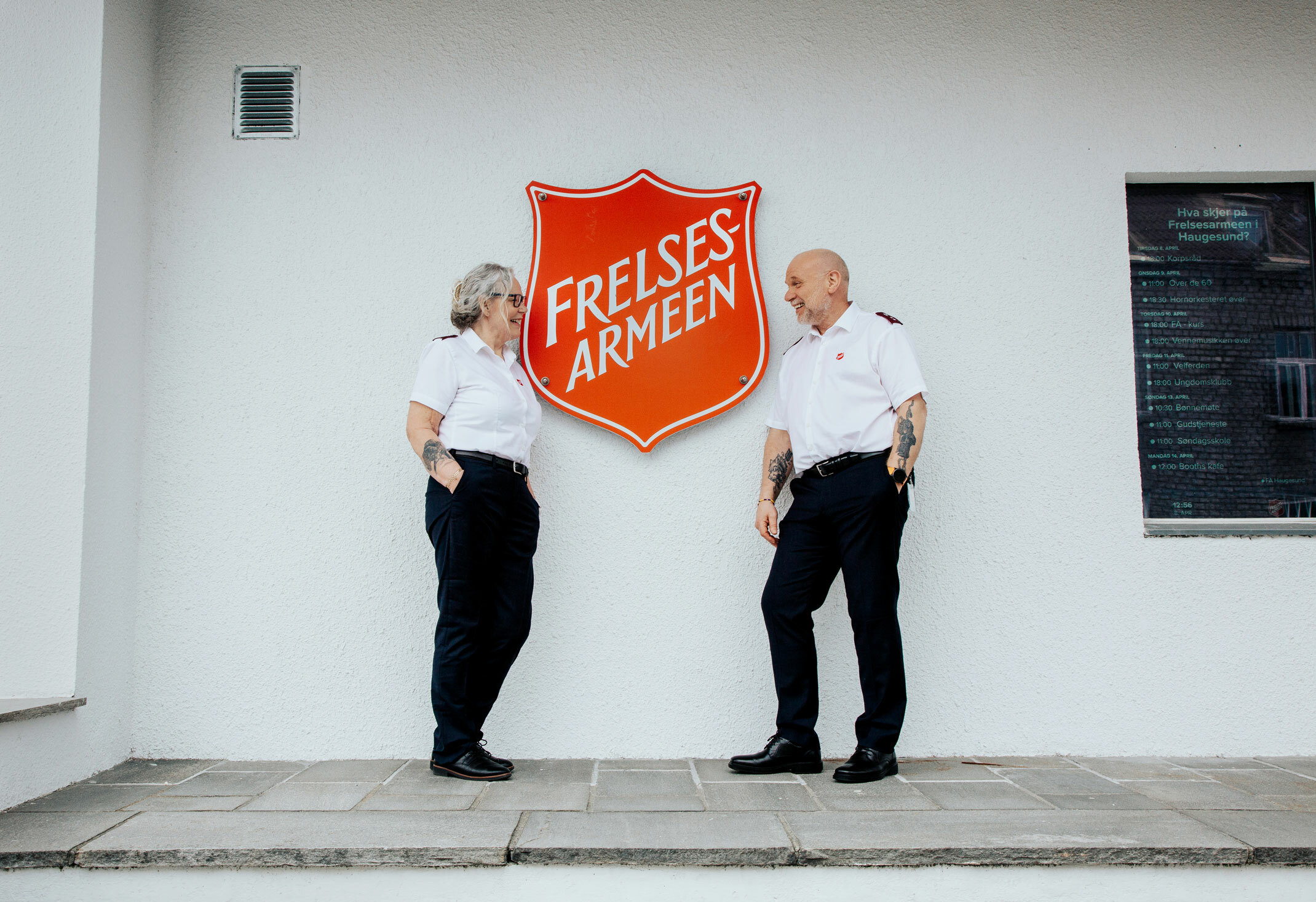 Foto: Kvinne og mann står på hver sin side av en diger, rød logo fra Frelsesarmeen. Den henger på en hvit murvegg.