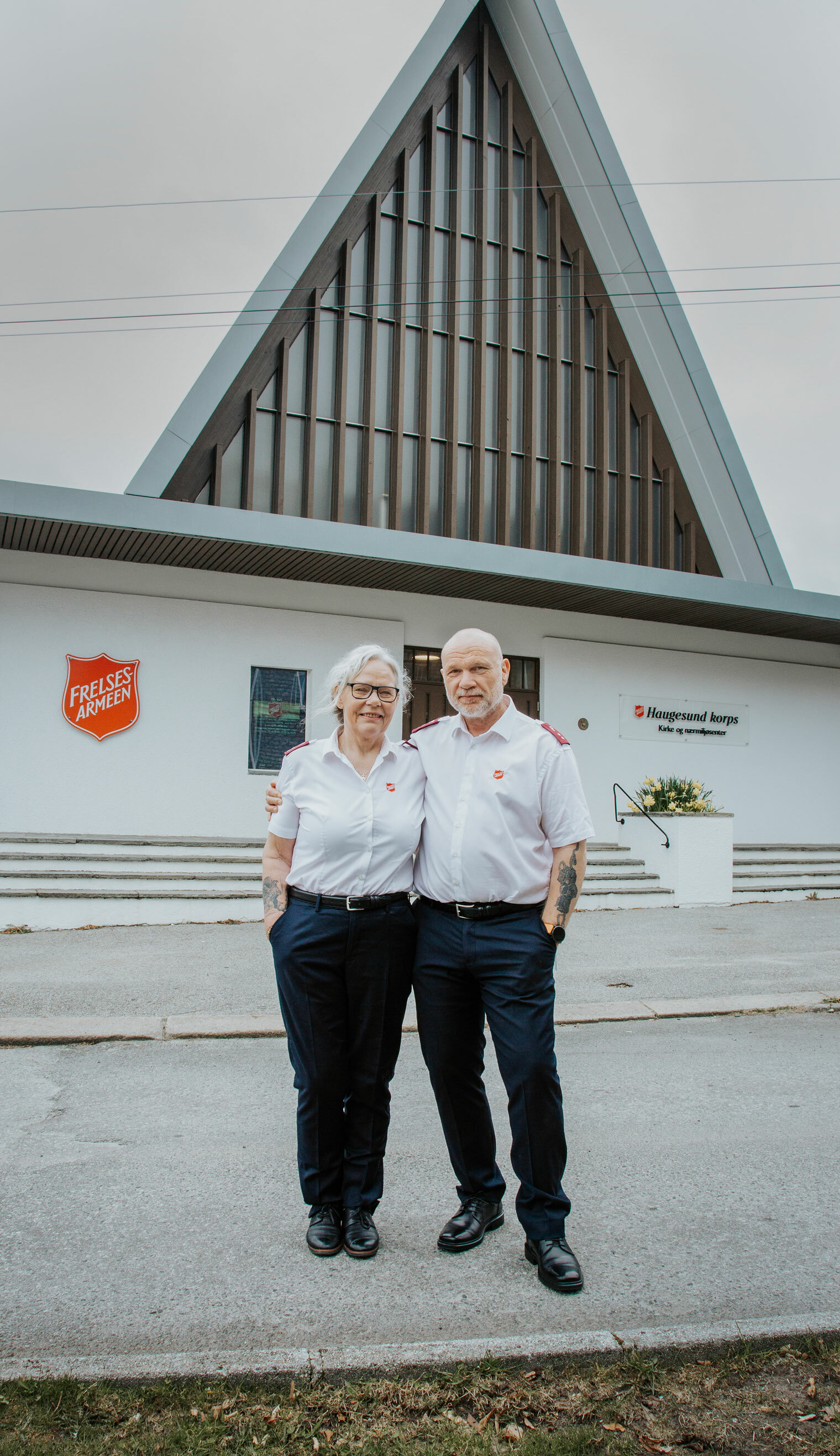 Foto: Middelaldrende kvinne og mann i uniform fra Frelsesarmeen poserer foran kirkebygget med spisst tak.