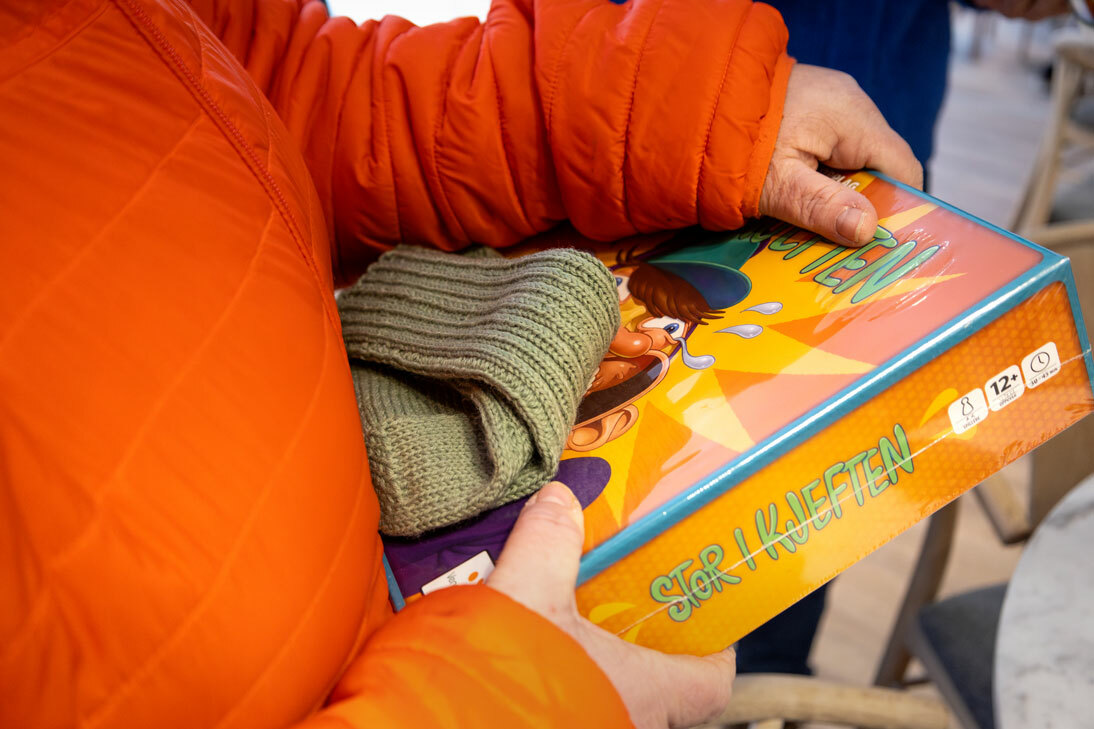 Foto: Kvinne i en oransje boblejakke bærer en eske med et spill. Oppå ligger et hjemmestrikkede ullstrømper.