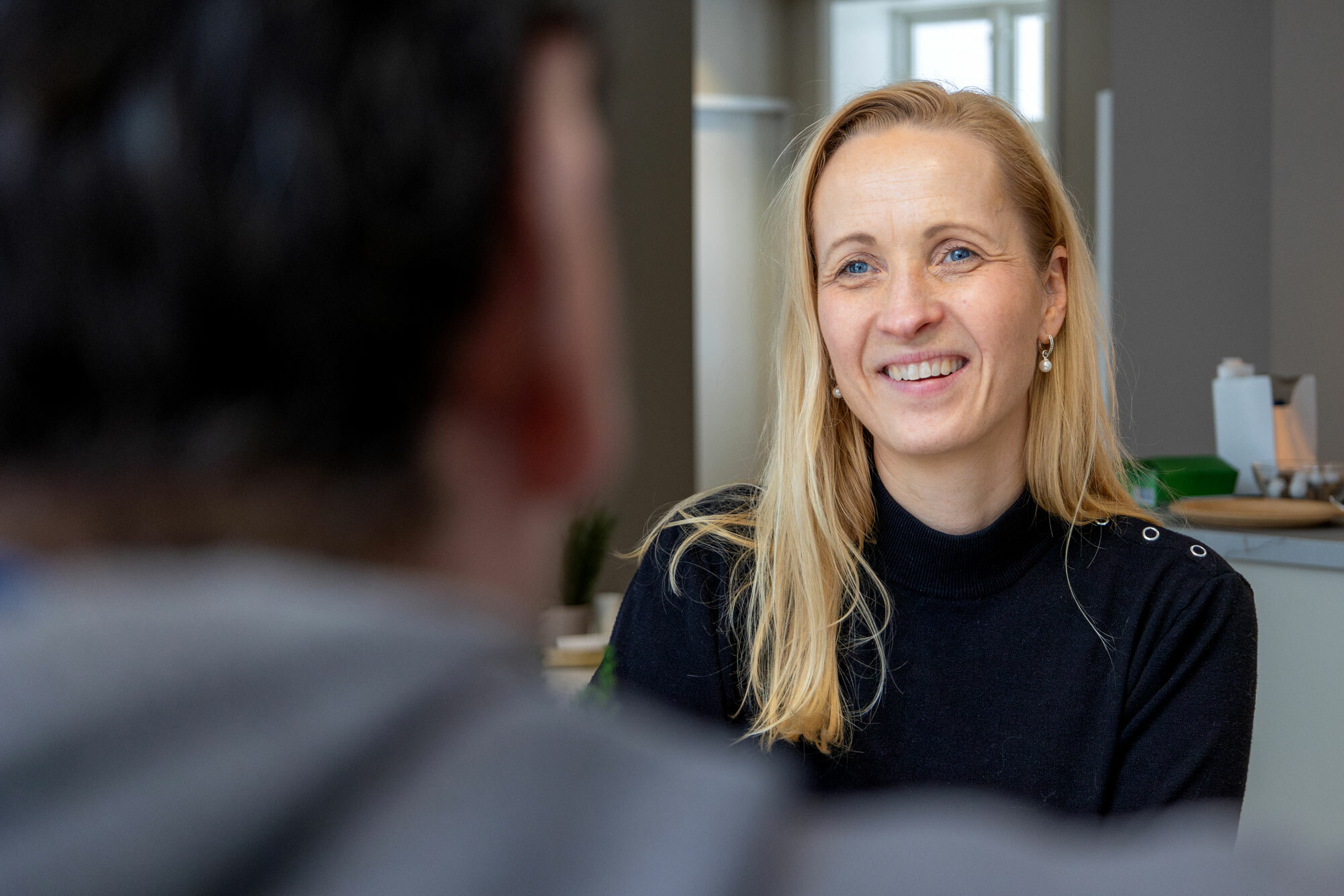 Foto: Kvinne i 30-årene med blondt hår snakker med en mann som sitter med ryggen til.