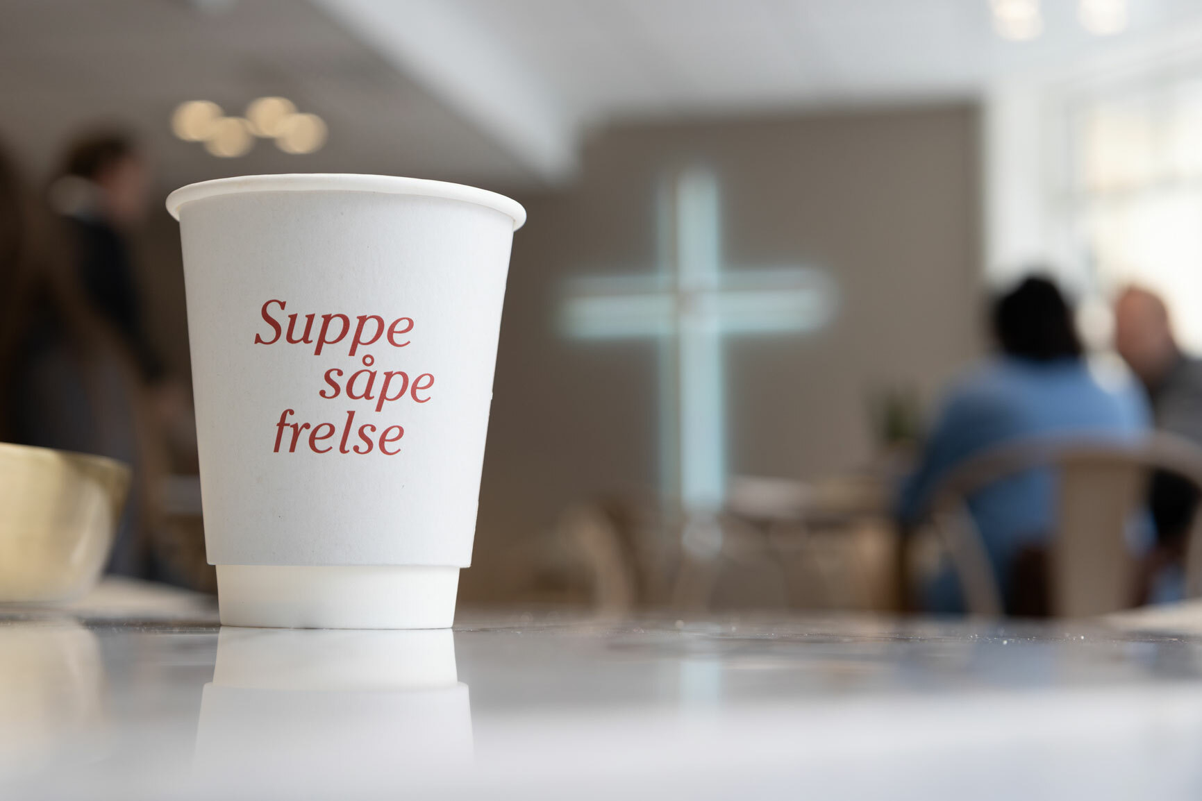 Foto: Et hvitt pappkrus står på et bord er i fokus i bildet. Bak i rommet ser vi at det sitter mennesker og snakker og vi ser et lysende kors på veggen.