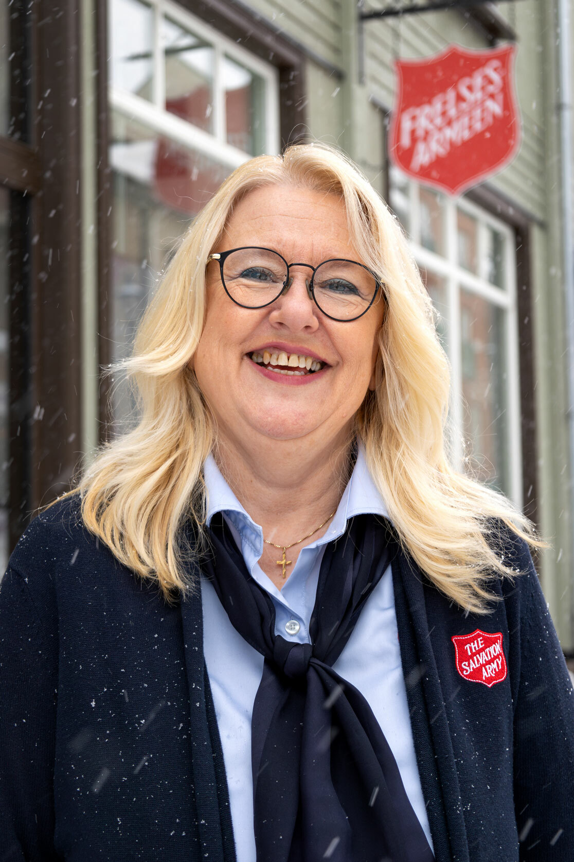Foto: Kvinne med halvlangt blondt hår og briller i klær fra Frelsesarmeen. Hun står utenfor et lokale med skilt inn til Frelsesarmeen.