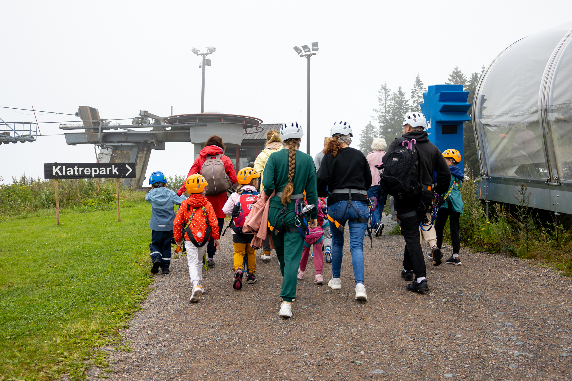 Foto: En gjeng med folk går bortover en grussti. Det står et skilt med klatrepark der og en stille skiheis på denne tiden av året. Litt tåke over grønne marker.