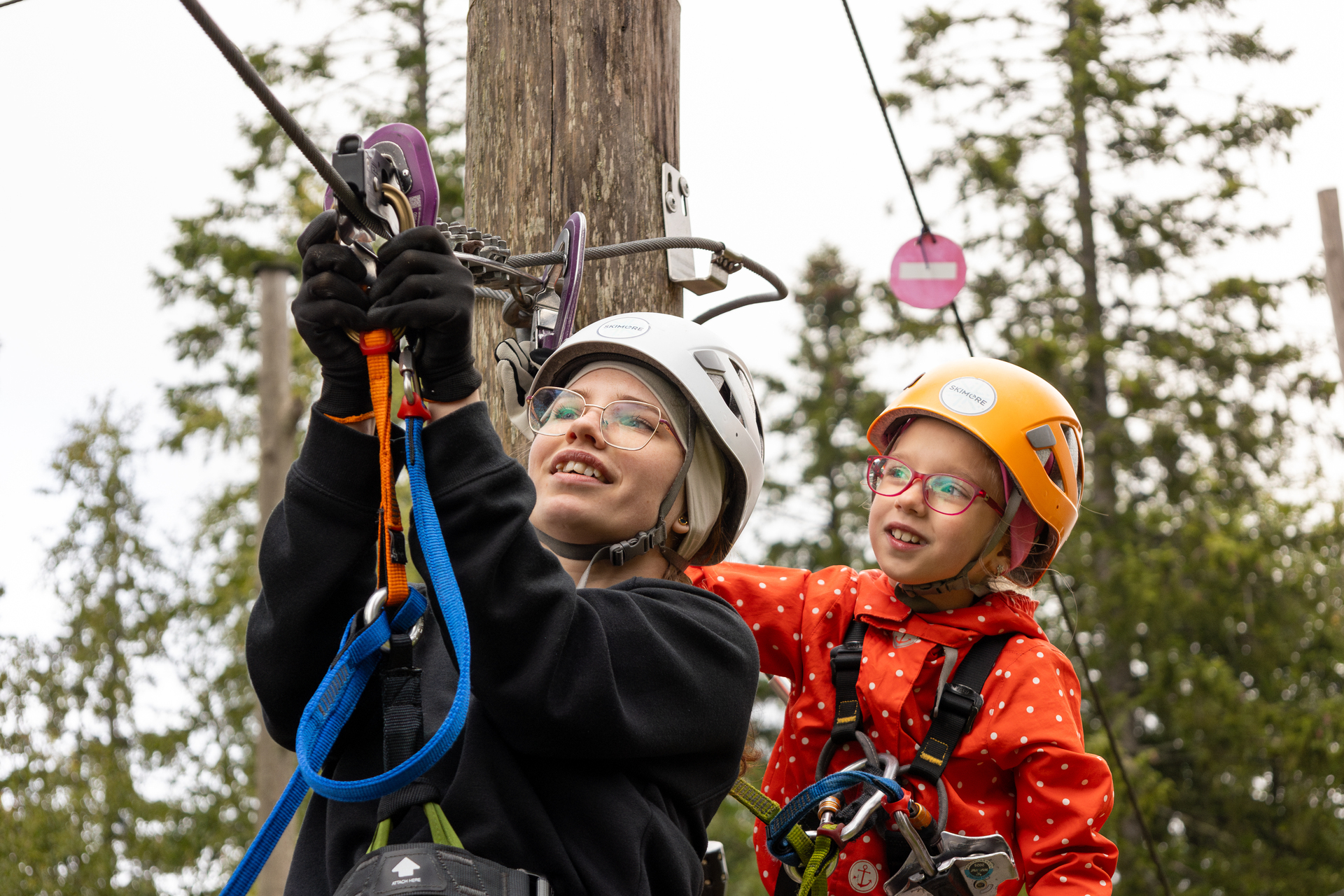 Foto: Mor og mindreårig datter med hjelmer på hodet fester eb karabin til en stålvaier ved et tre.