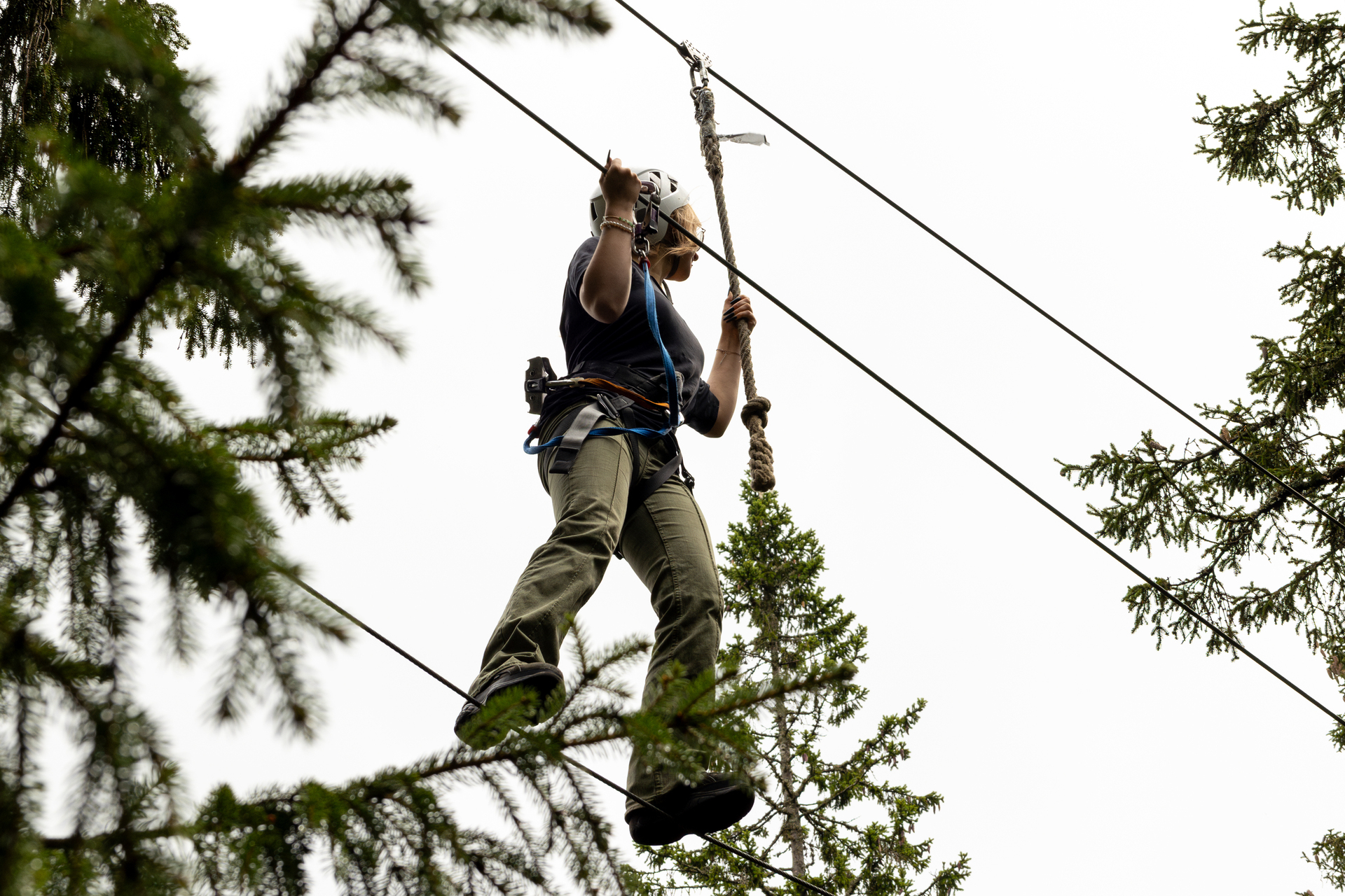Foto: Kvinne går på en line høyt oppe blant trærne. Hun er godt sikret med sæle og et tau.