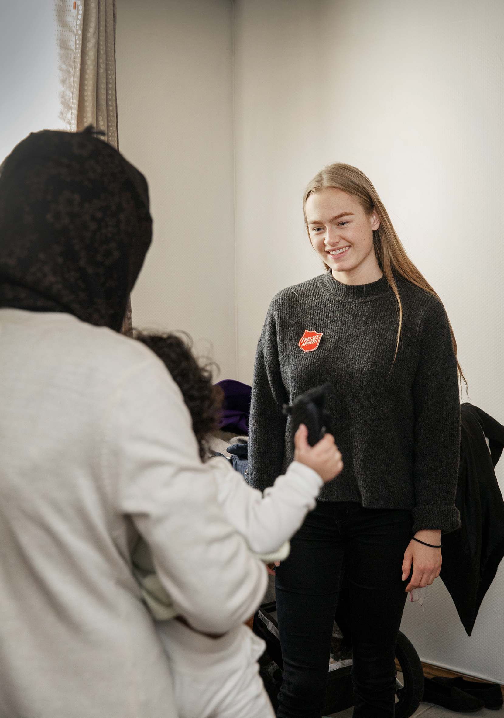 Foto: Ung kvinne med langt hår og logo fra Frelsesarmeen på genseren snakker med en dame med svart hijab som står med ryggen til kamera. Hun holder et lite barn med mørke krøller.