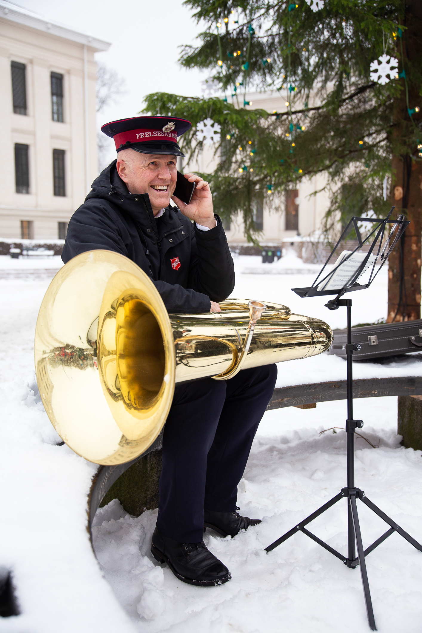 Mann mørk uniform og hatt med stor tuba på fanget. Snakker i mobilen.