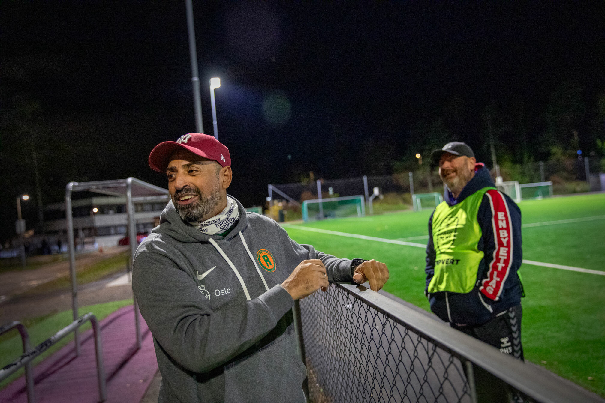 Foto: To menn står på hver sin side av et gjerde rundt en fotballbane i flombelysningen. Det er kveld og irrgrønn bane.