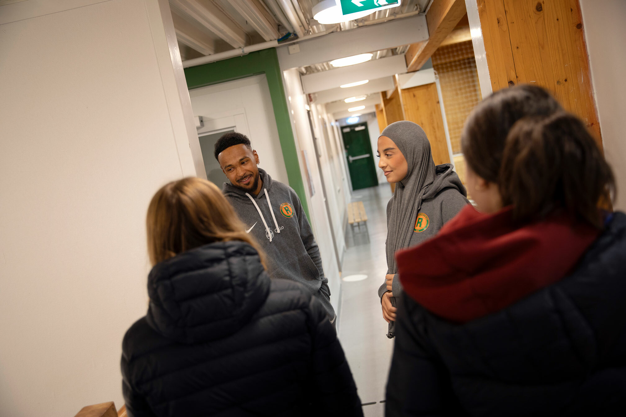 Foto: To leder i grå hettegensere med logo fra Romsås IL står i en gang. Vi ser ryggen til to barn som kommer imot dem.