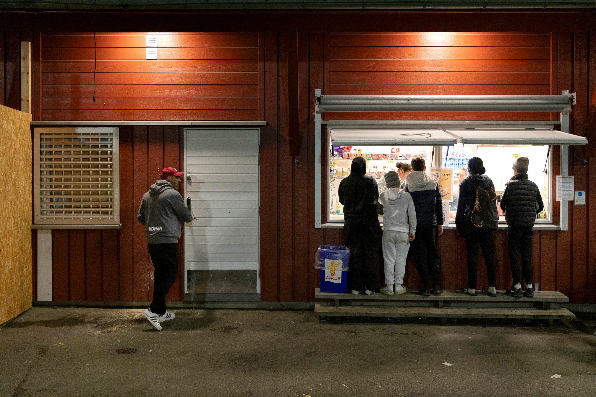 Foto: Bilde av en mann som går mot en dør. Ved siden av er en kiosk gjennom en åpning i veggen. Mange ungdommer står i kø der.