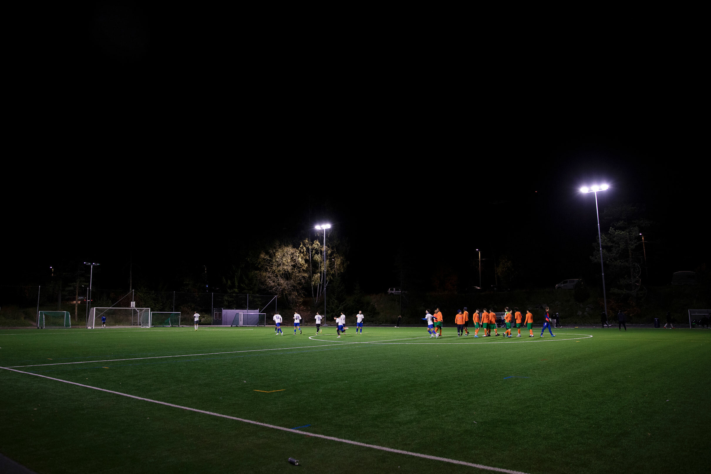 Foto: På en stor gressbane ser vi to lag med spillere i hvite og røde drakter. Store lykter lyser opp gressmatta i mørket.