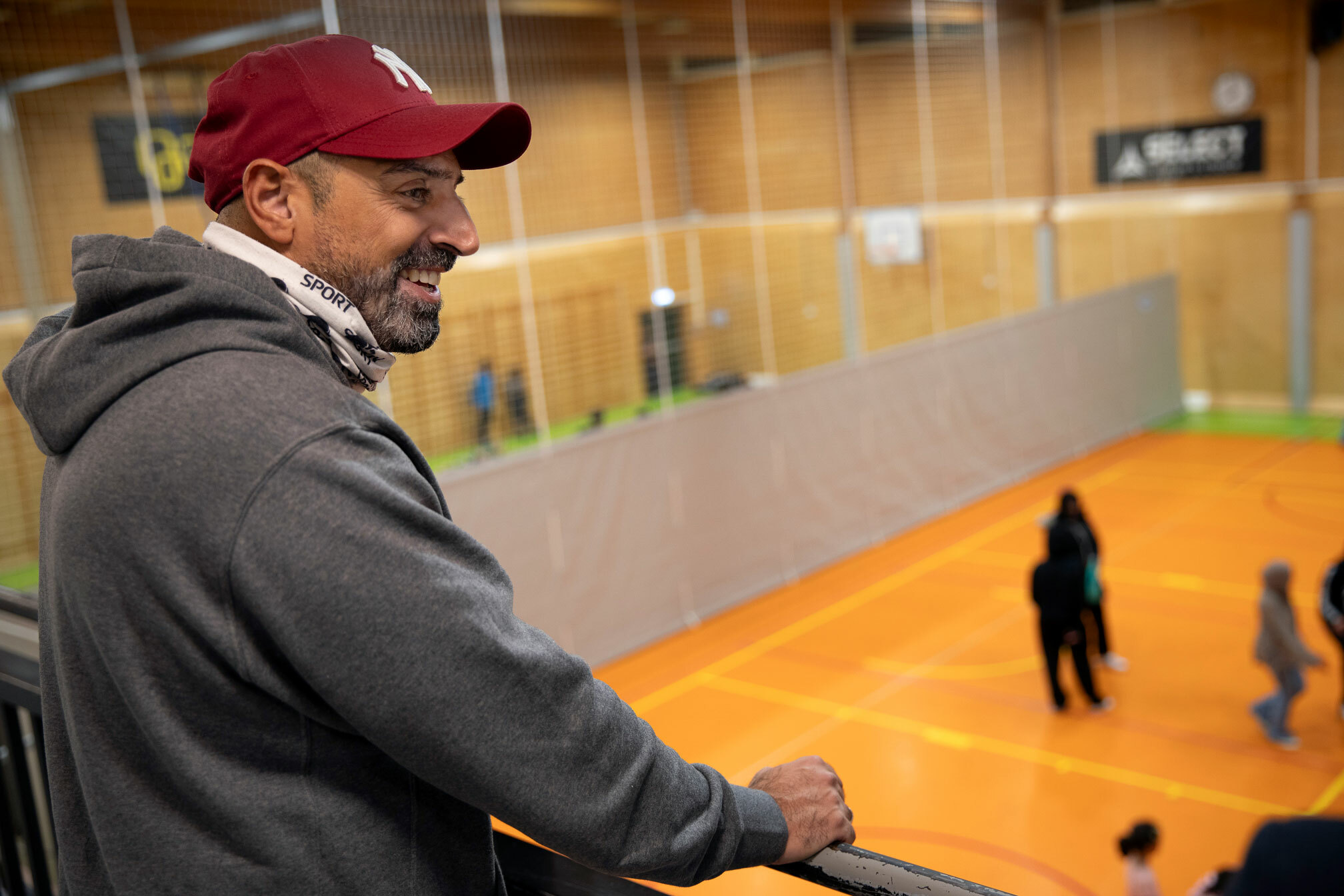 Foto: Mann i treningstøy og caps står oppe på en tribune og ser barn leke i en innendørshall.