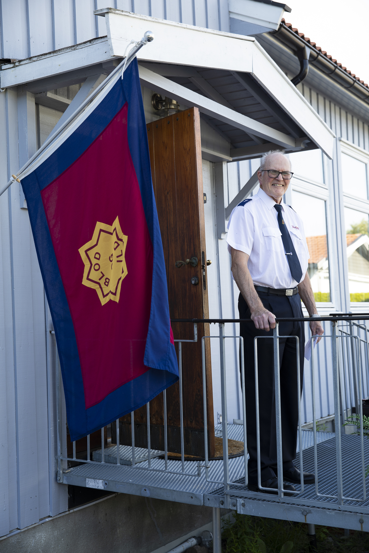 Eldre mann i kortermet Frelsesarméuniform står ute på trappa foran et forsamlingslokale. Frelsesarmeens flagg er satt opp ved inngangsdøren