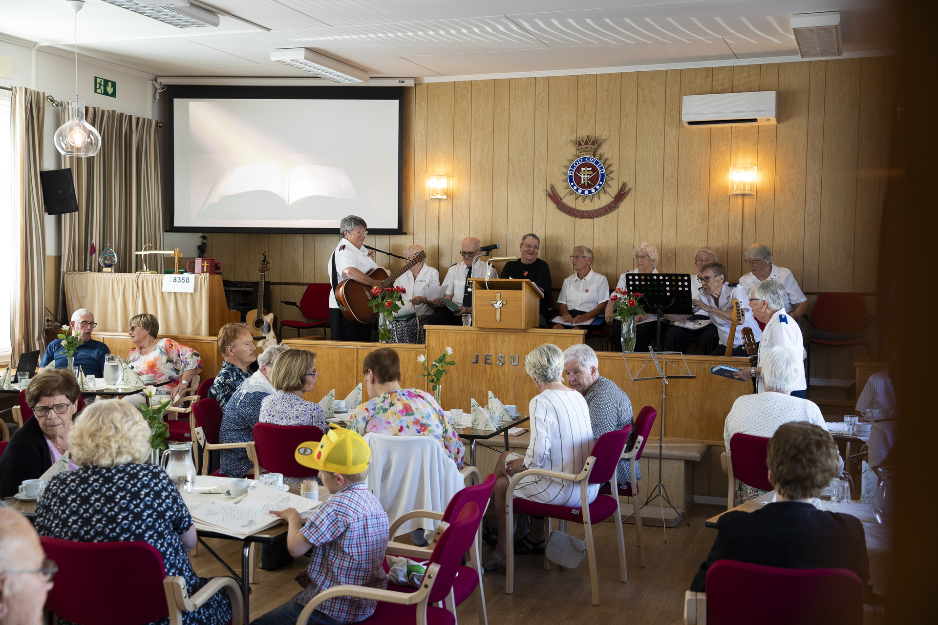 Folk sitter rundt små bord i et foramlingslokale.