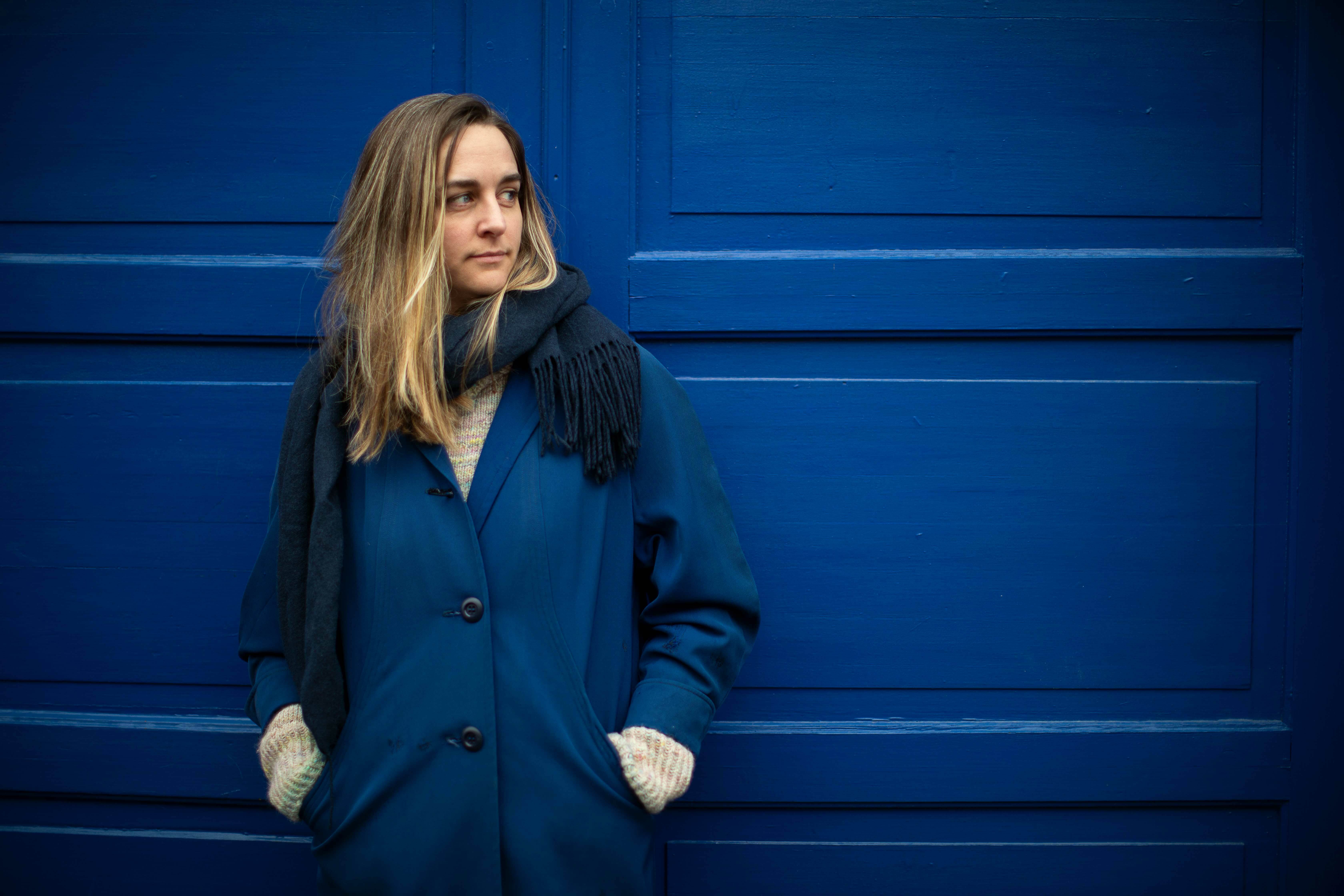 Kvinne med halvlangt hår og mørkt skjerf står lent mot en blå dør.