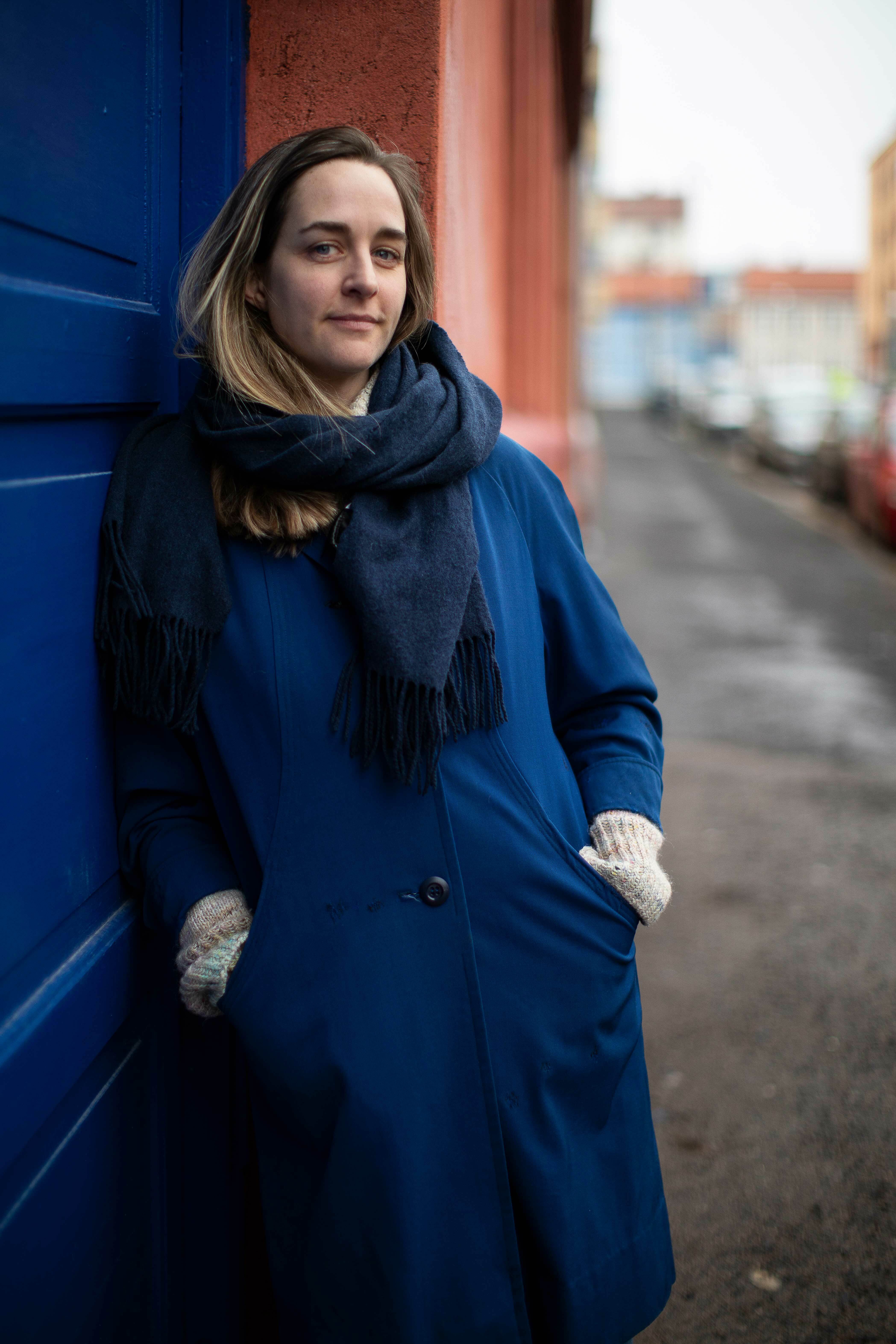 Kvinne med halvlangt hår og mørkt skjerf står lent mot en blå dør.