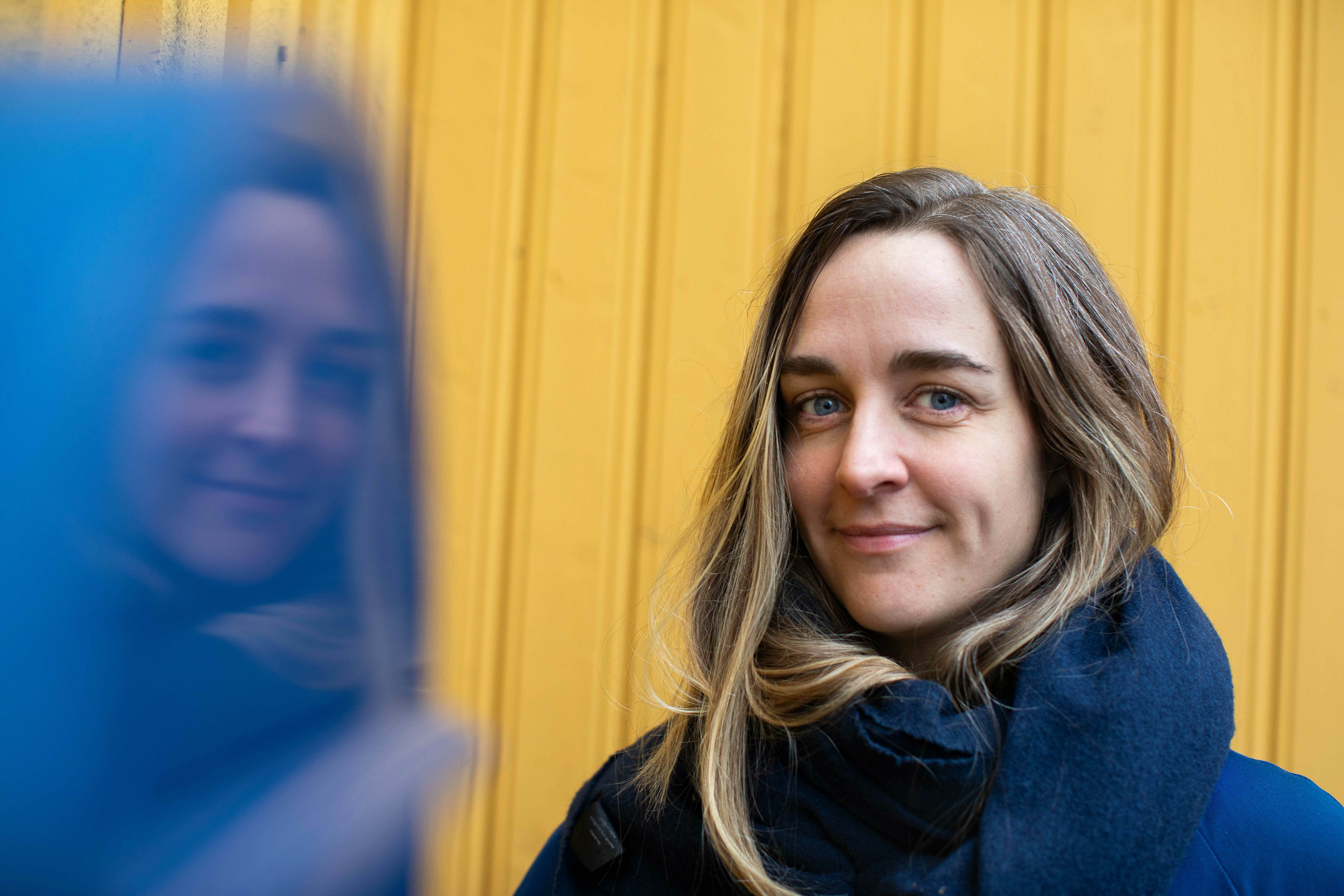 Kvinne med halvlangt hår med blå kåpe mot en gul vegg.