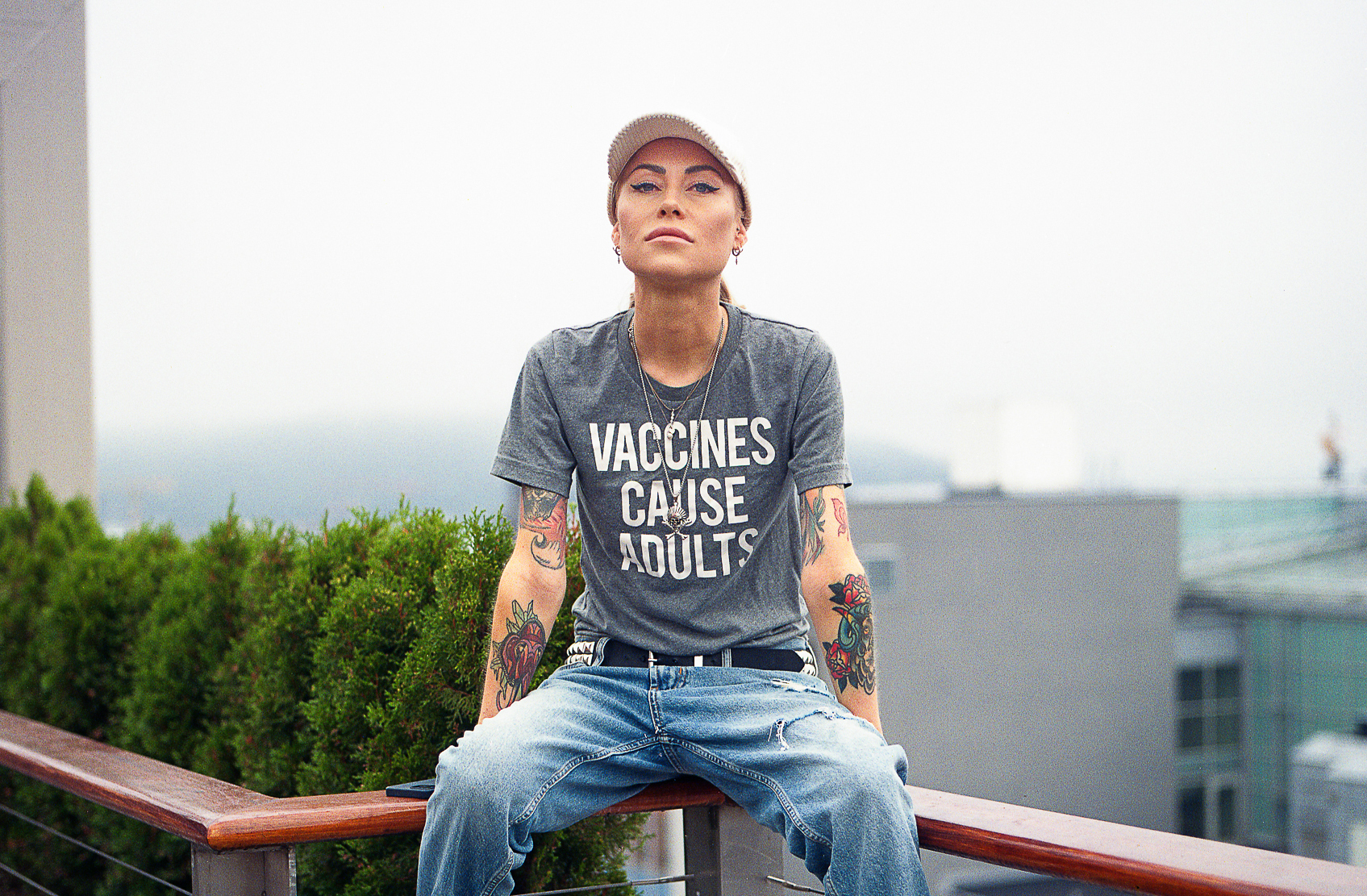 Kvinne i 30-årene sitter på et rekkverk i et høyt hus. Hun har caps, T-skjorte og dongeribukse på seg. Armene er fulle av fargerike tatoveringer.