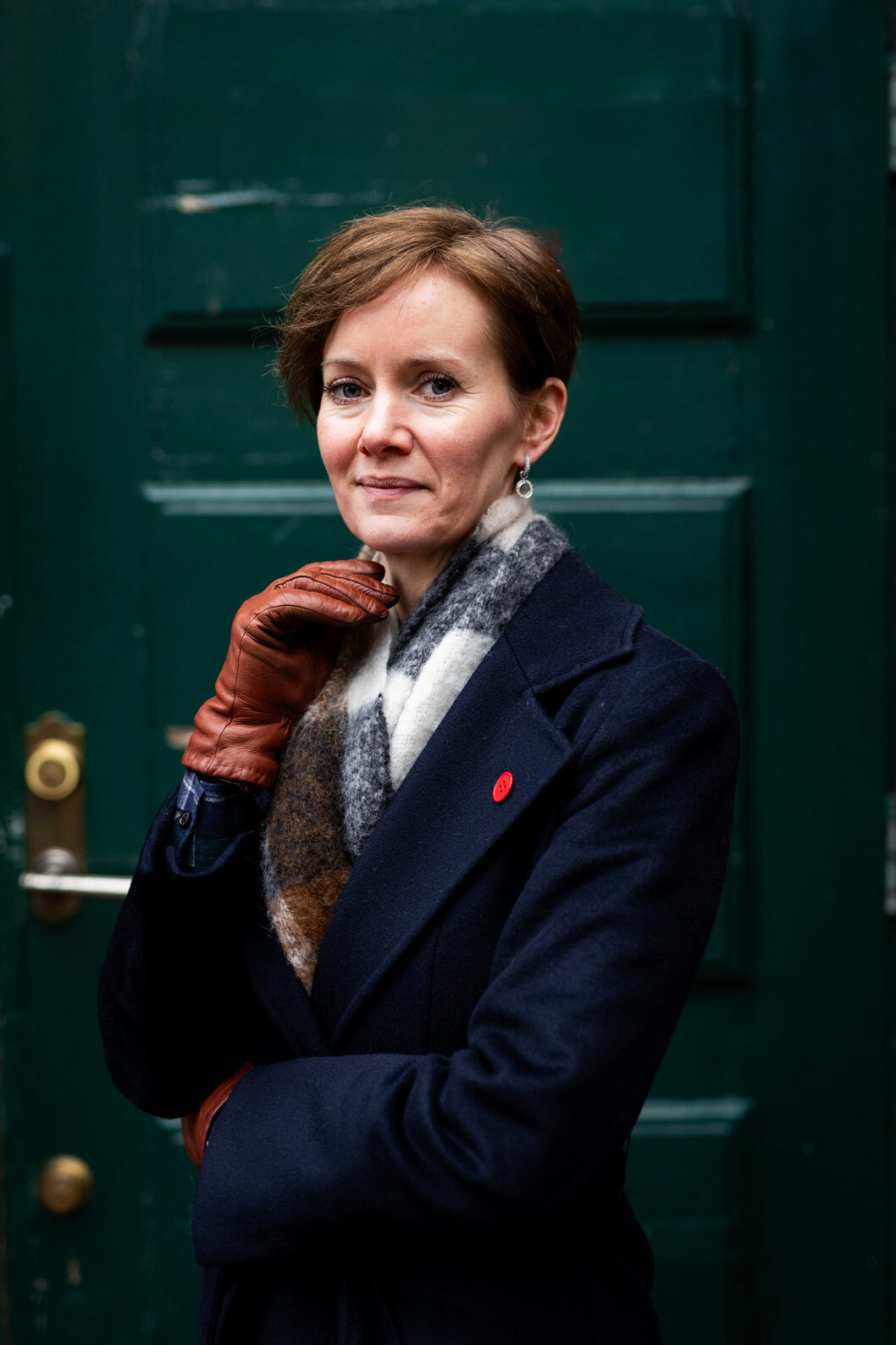 Foto: Kvinne med kort hår står foran en grønn dør. Hun har på seg kåpe og skjerf, og holder høyre håne med skinnhanske opp mot haken.