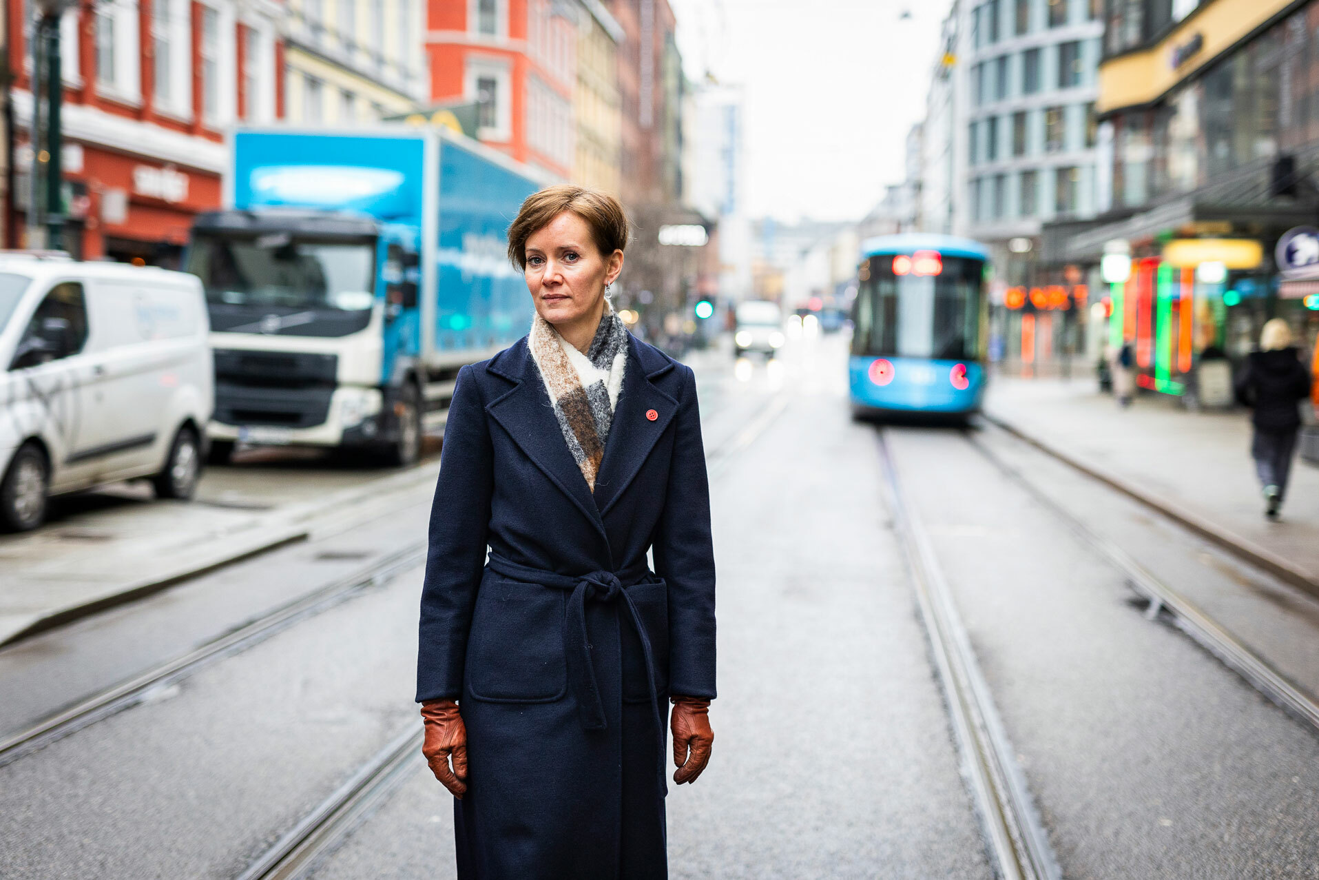 Foto: Kvinne med kort, rødlig hår står midt i en bygate i Oslo med trikkespor. Det er tidlig vår og hun har på seg varm kåpe, hansker og skjerf.