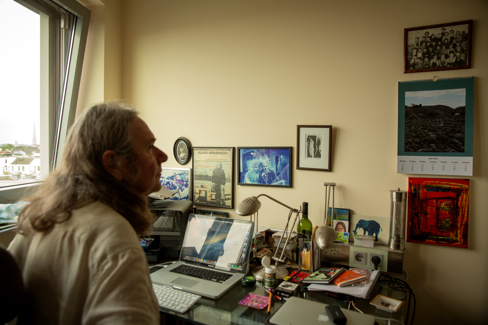 Foto: Mann med halvlangt, brunt hår sitter inne på et lite, gulmalt rom med skrivepult. På veggen henger flere bilder og en kalender.