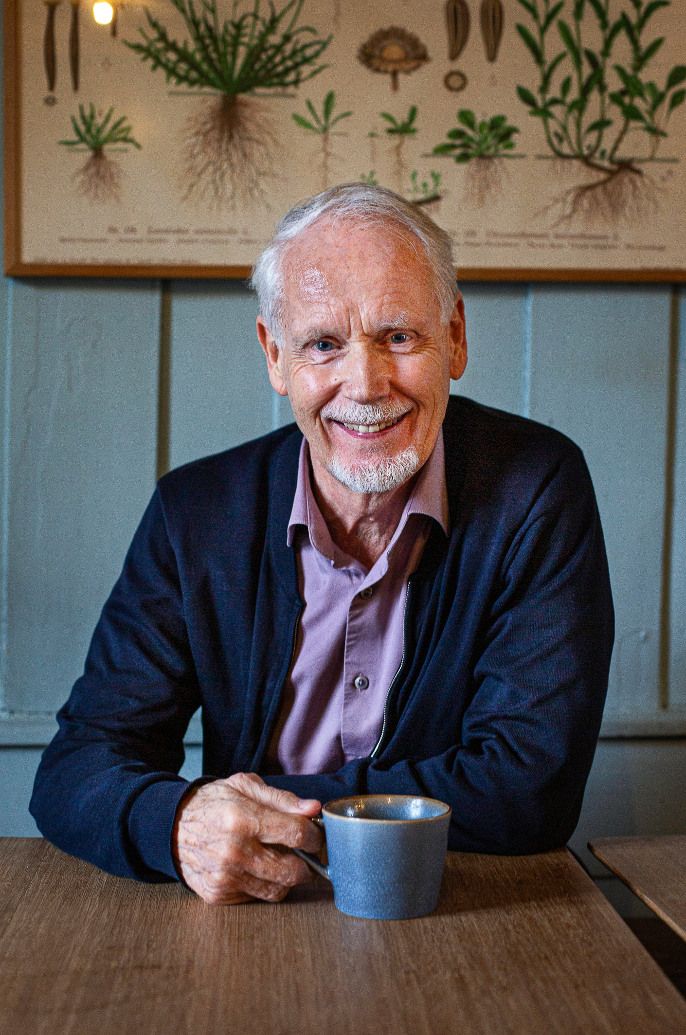 Foto: Mann med kopp i hånden. Han sitter ved et kafebord og smiler mot kamera. Han har grått hår og skjegg.