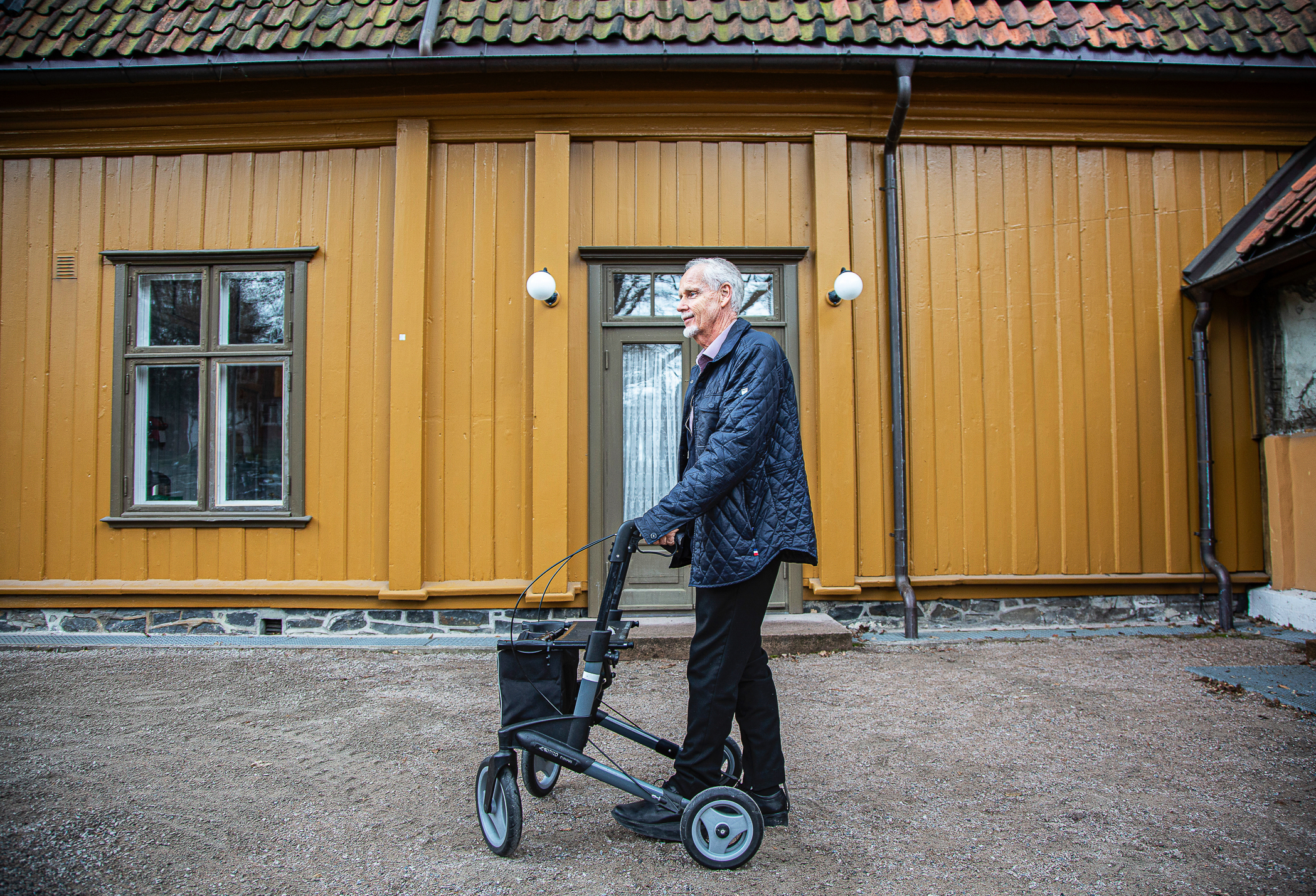Foto: Eldre herre i mørke klær går forbi et gammelt, okergult hus med sin rullator.