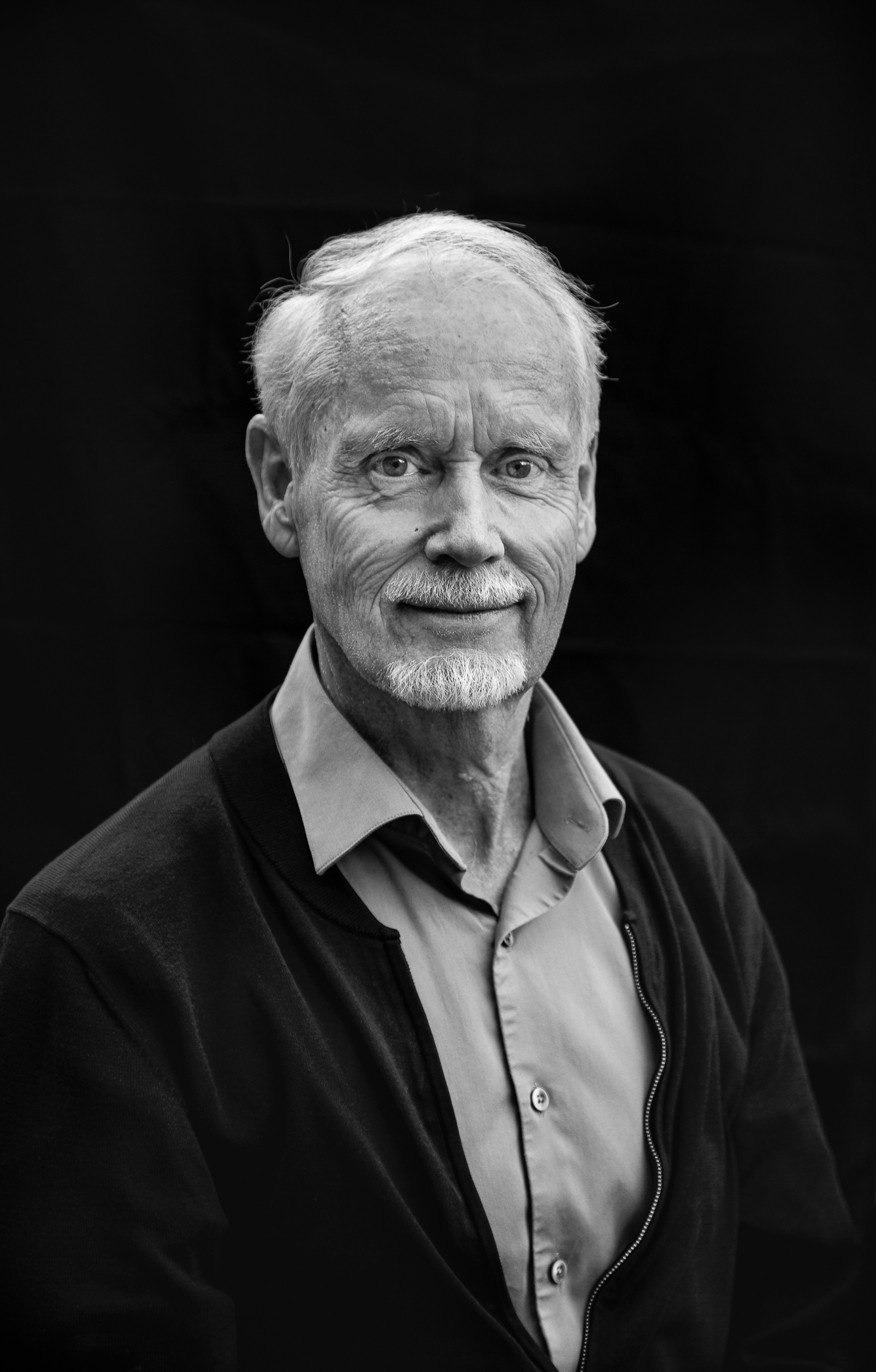 Foto: Portrett i sort/hvitt av en eldre herre med et direkte, fast blikk og et lite smil. Han har grått hår og skjegg.
