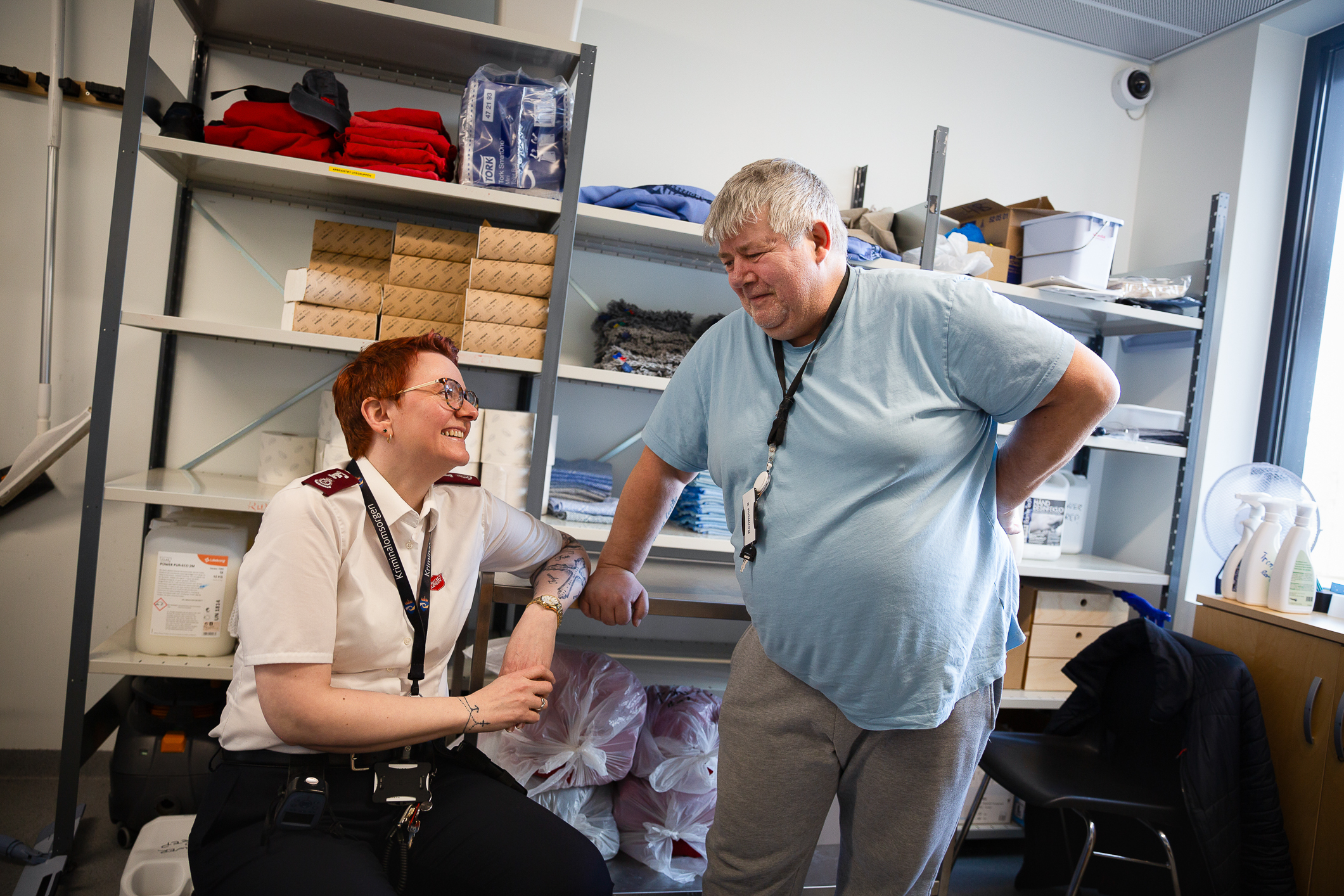 Ung kvinne med kortermet frelsesarme-skjorte sitter på ei bøtte på et lagerrom og prater med en middelaldrende mann med lys blå t-skjorte. Kvinnen har dyp rød farget hår. Begge har nøkkelkort hengende i en stropp rundt halsen.