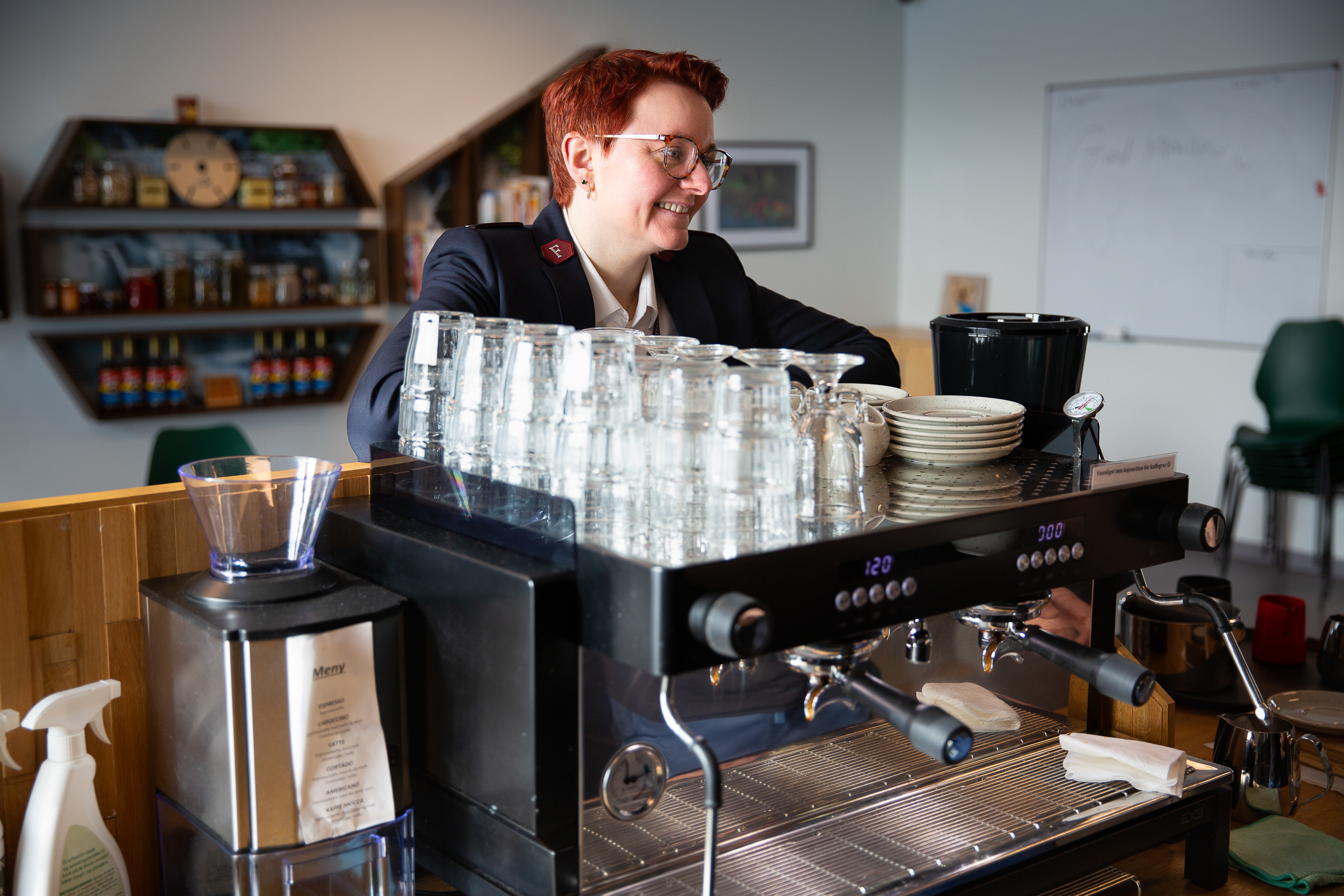 En ung kvinne med frelsesarméuniform og kort, hennarødt hår og briller står og venter ved disken på en kafé. Bak disken ser vi en proff kaffemaskin til å lage baristakaffe med.