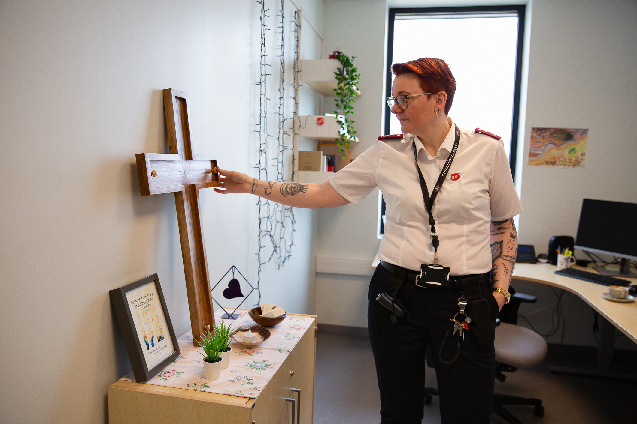 Ung kvinne med frelsesarmeuniform, kort hår og briller står og holder et trekors i et kontorrom. Hun har kortermet skjorte, tatoveringer på armene og håret har en dyp rød farge