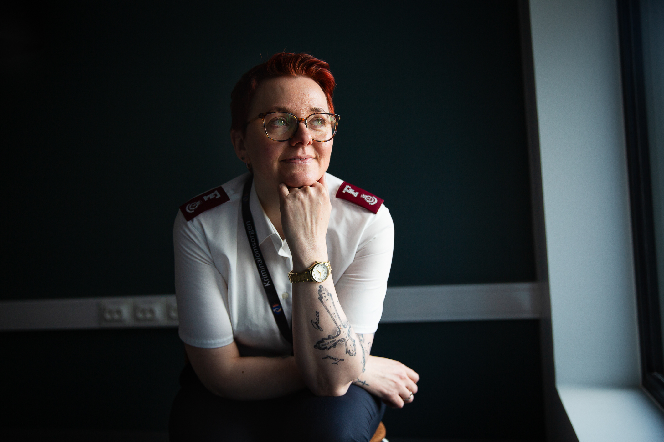 Ung kvinne med frelsesarmeuniform, kort hår og briller. Hun sitter i et mørkt rom med lys fra vinduet. Hun har briller og tatoveringer på armen håret har en dyp rød farge