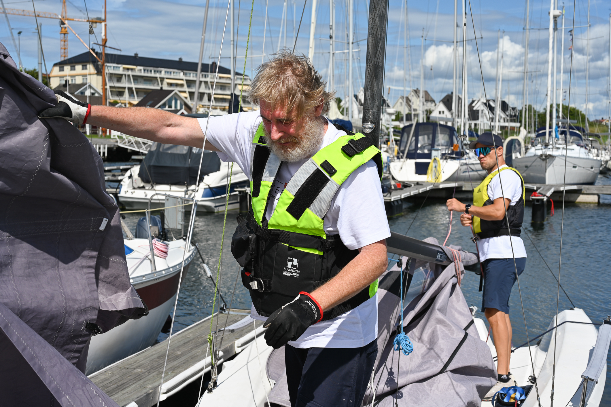 To menn i en seilbåt som ligger til kai. De rigger ned seil.