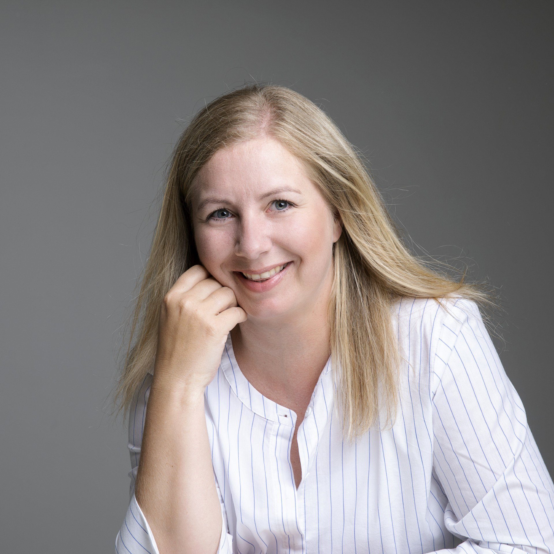 Janette Røseth sitter og smiler mot kamera. Hun lener seg på den ene armen, hun har på seg lys stripete skjorte og bakgrunnen er grå.