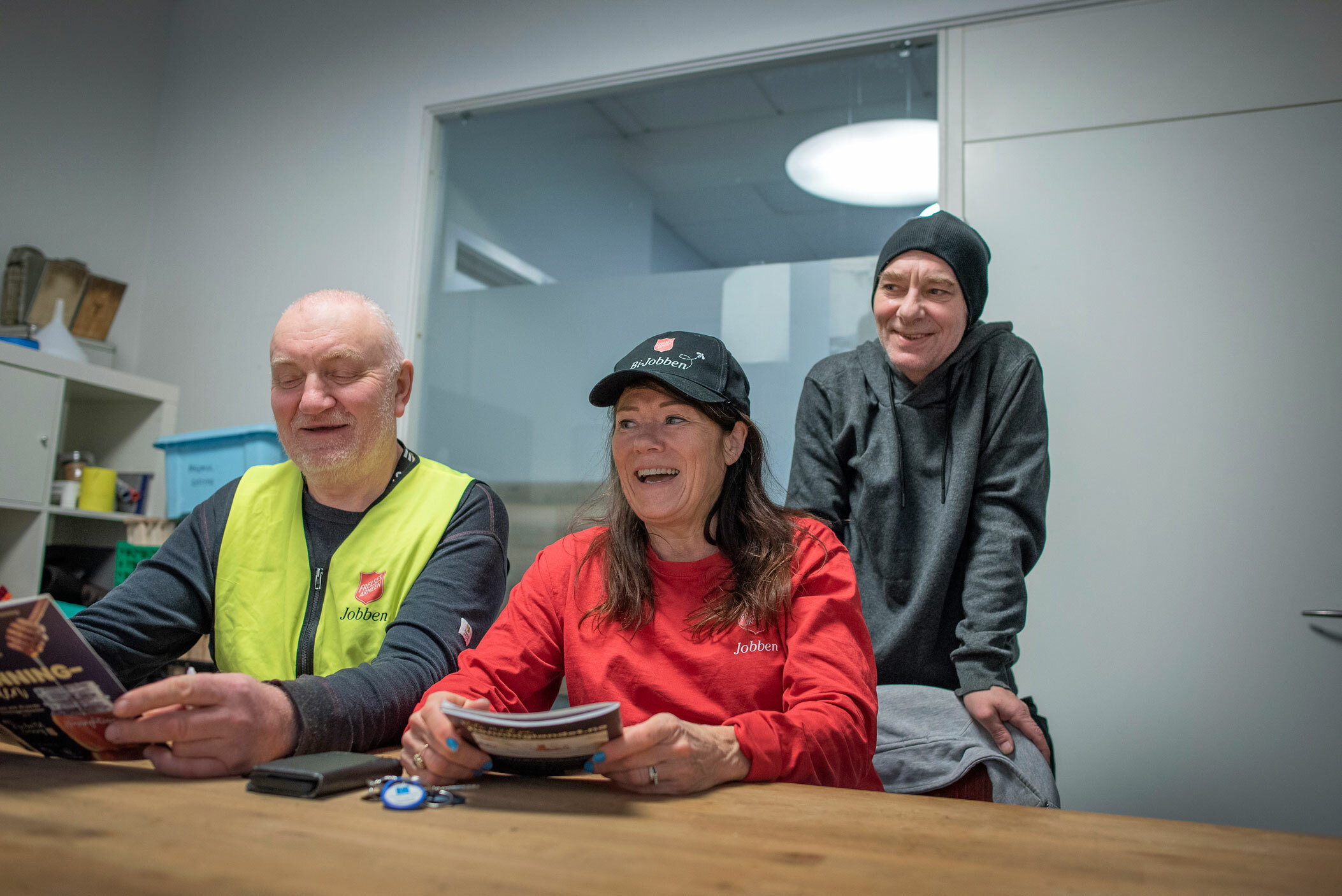 Foto: En kvinne med svart caps over langt, mørkt hår har på seg en rød genser med logo fra Frelsesarmeen. Hun sitter ved et bord sammen med en kar i gul refleksvest fra Jobben. Bak den står en mann i grå klær med lue på.
