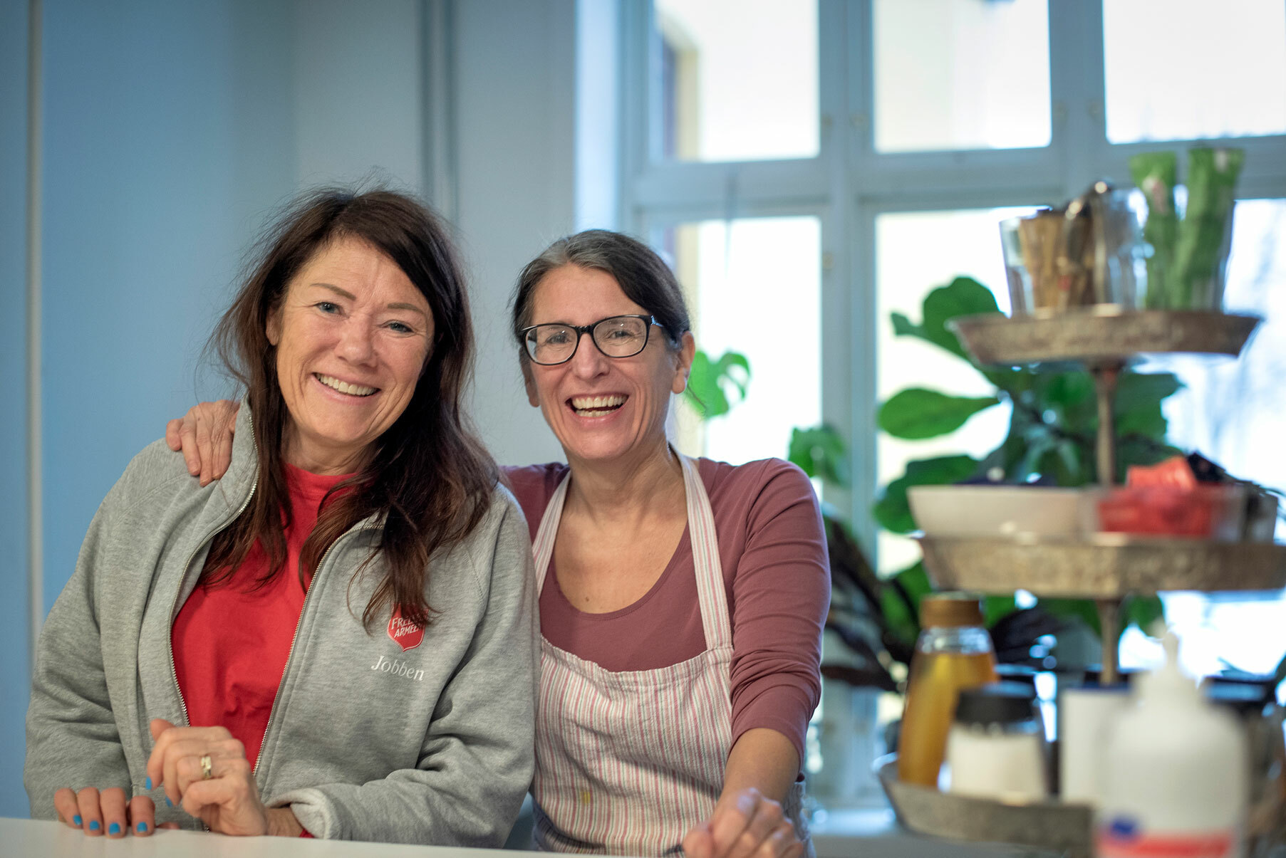 Foto: To kvinner i 50-årene står tett sammen bak en disk. De smiler mot kamera. Den ene har briller og et forkle på seg, den andre både grå hettegenser og rød genser fra Frelsesarmeen.