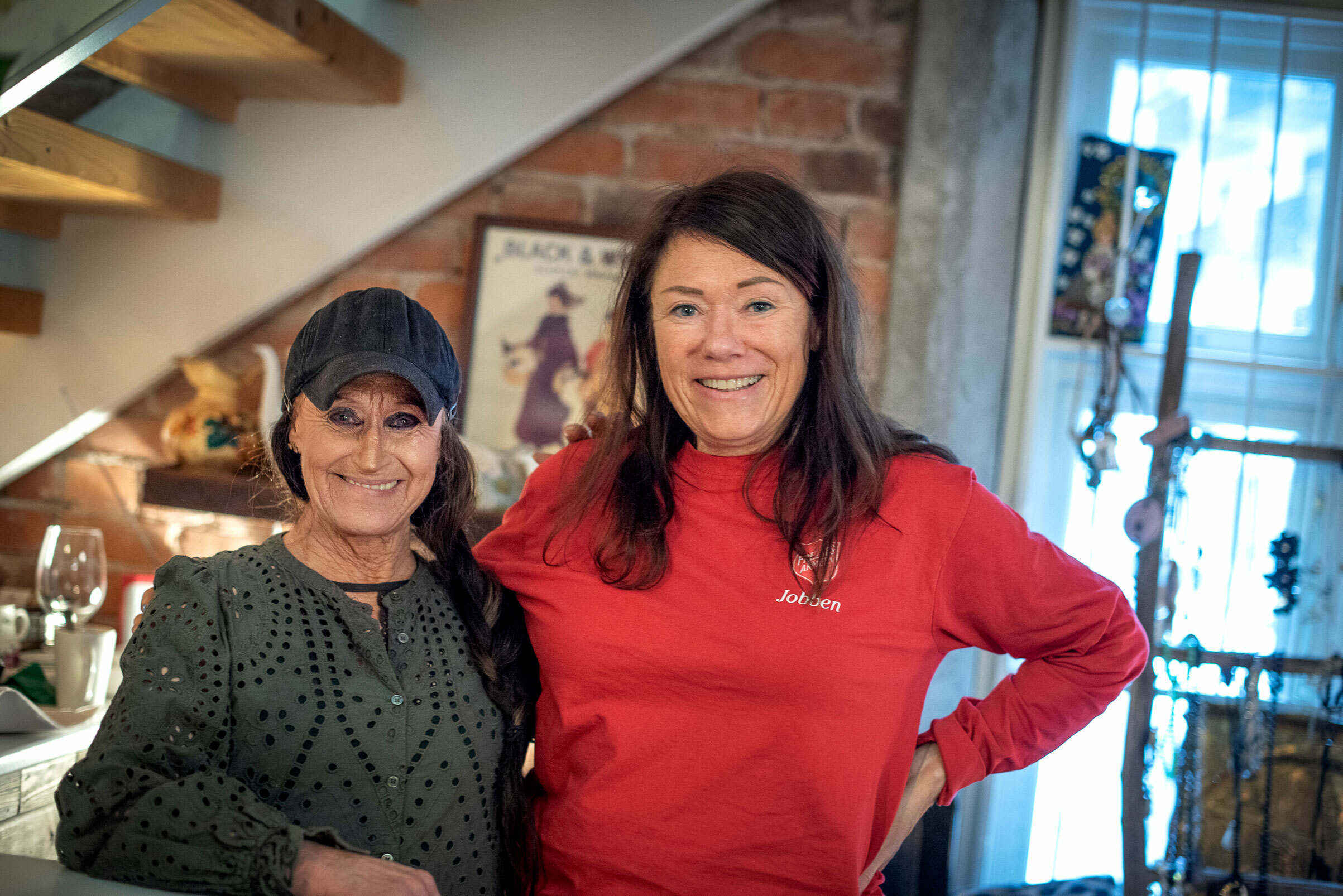 Foto: To kvinner står ved siden av hverandre i et butikklokale under ei trapp. Begge er i slutten av 50-¨rene og ei er kledd i grønn penbluse og ei i rød genser med logoen til Frelsesarmeen på.