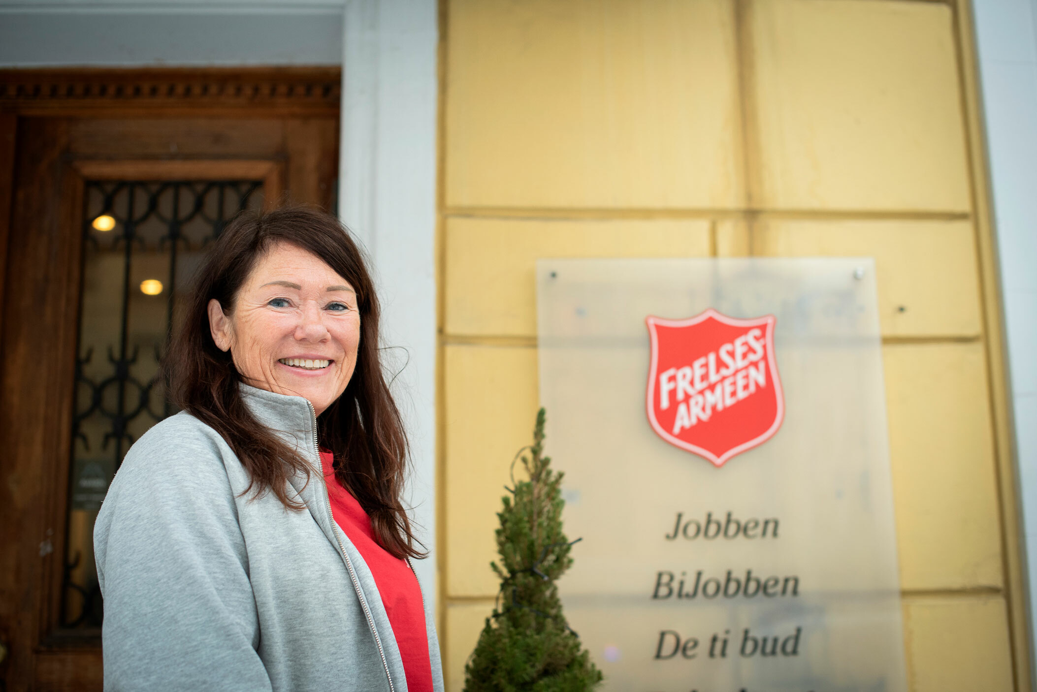 Foto: Kvinne med mørkt, langt hår står foran inngangsdøra til Jobben. På en gul murvegg henger skiltet med Frelsesarmeens røde logo.