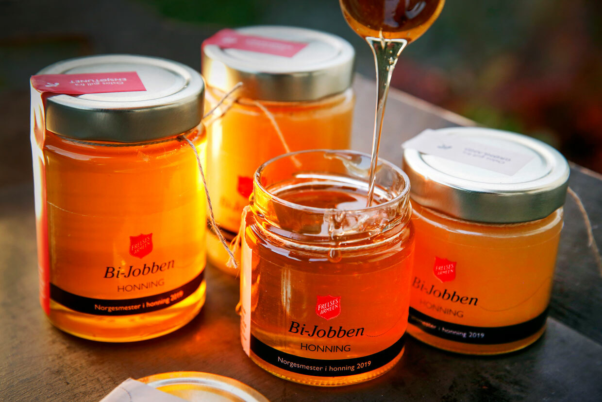 Foto: Tre fylte honningglass med lokk på står på et bord. Det helles honning i det fjerde. På etiketten er det logo fra Frelsesarmeen og det står Bi-Jobben på.