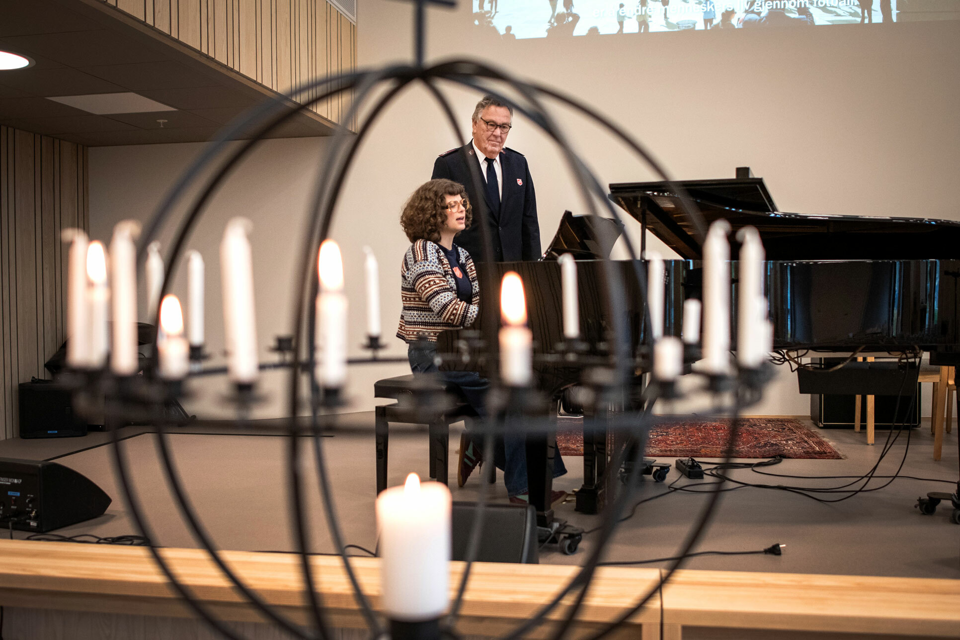 Foto: Mann i uniform står ved siden av et piano i et kirkerom hvor den unge kvinnen sitter og spiller.