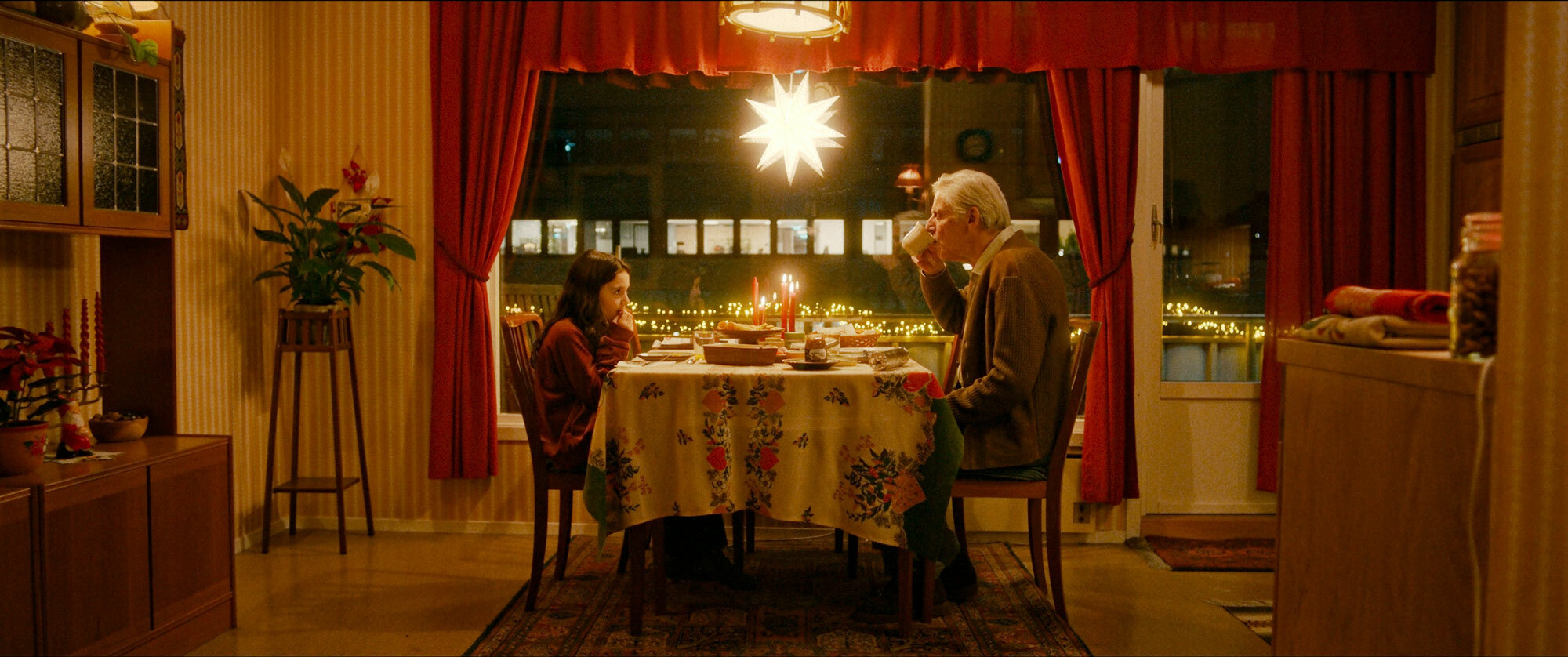 Foto: FRa en filmscene, så det er ekstra bredt bilde. En gammel mann med grått hår drikker fra en kopp. Han sitter i en stue som er innredet med røde gardiner og en julestjerne i vinduet. Stilen er fra 1960-tallet. Han sitter ved et spisebord der en liten jente med mørkt hår sitter på en stol. Bordet er dekket med lys og mar.