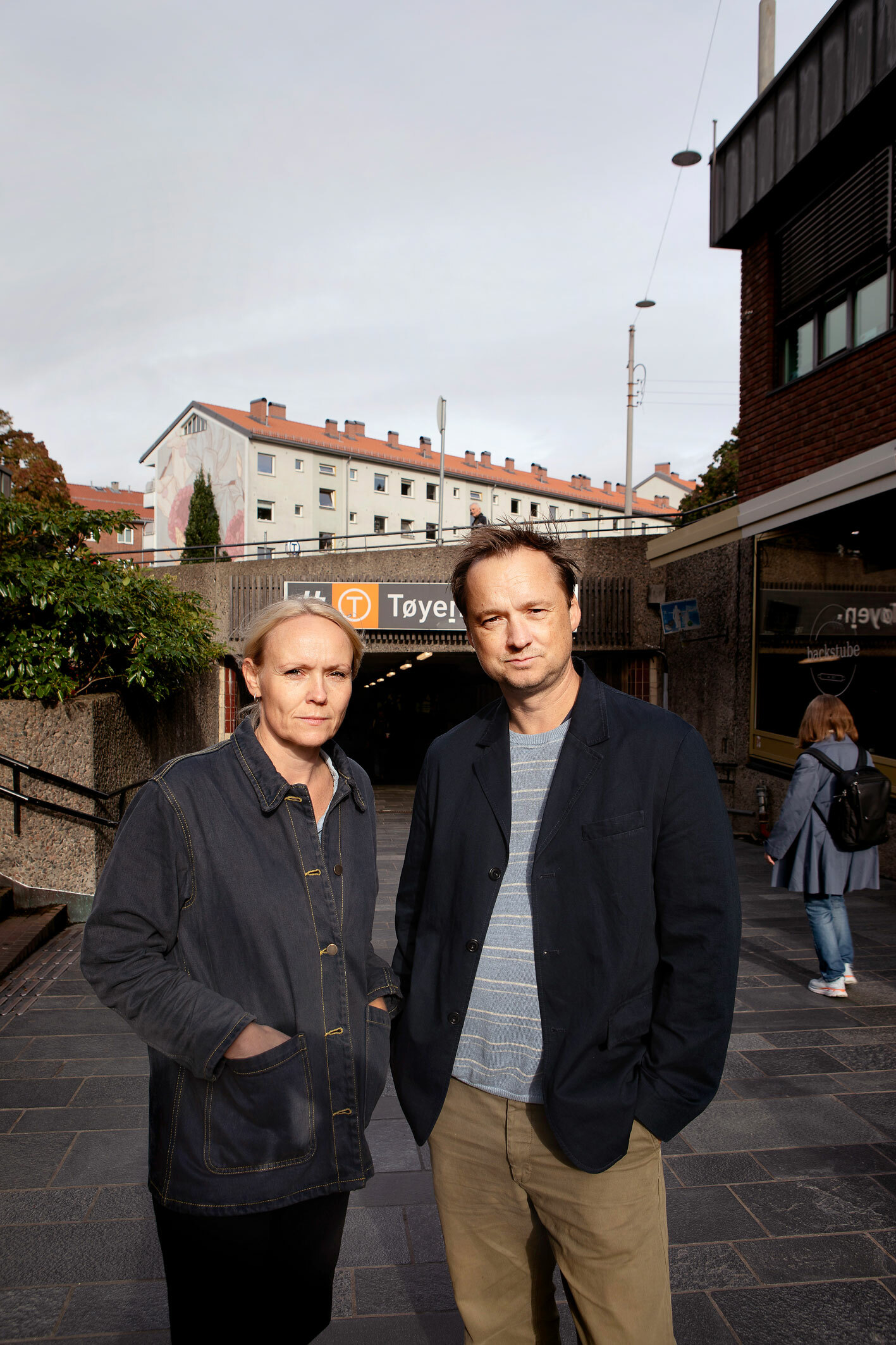 Foto: Foran inngangen til Tøyen T-banestasjon står den samme mannen i mellomblå genser og damen med blondt hår.