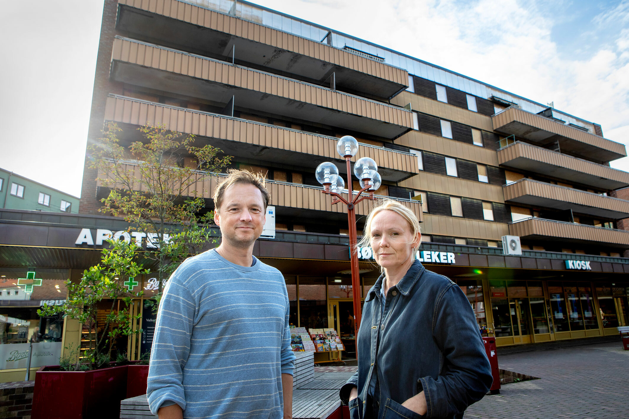 Foto: Utenfor en sliten blokk i Oslo står en mann i 40-årene med mørkeblondt hår i en mellomblå genser med striper. Ved siden av ham står en jevngammel, blond kvinne i dongerijakke.