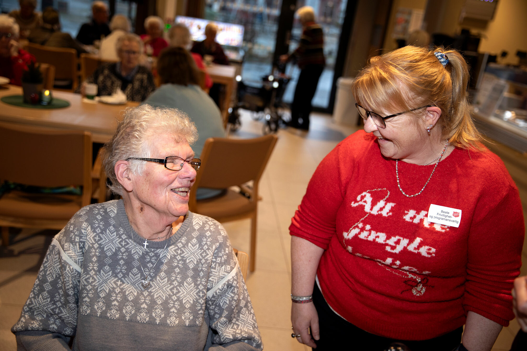 Foto: To damer snakker med hverandre, hun til venstre er eldst og sitter. Hun andre står og har på seg en rød julegenser. Begge har briller.