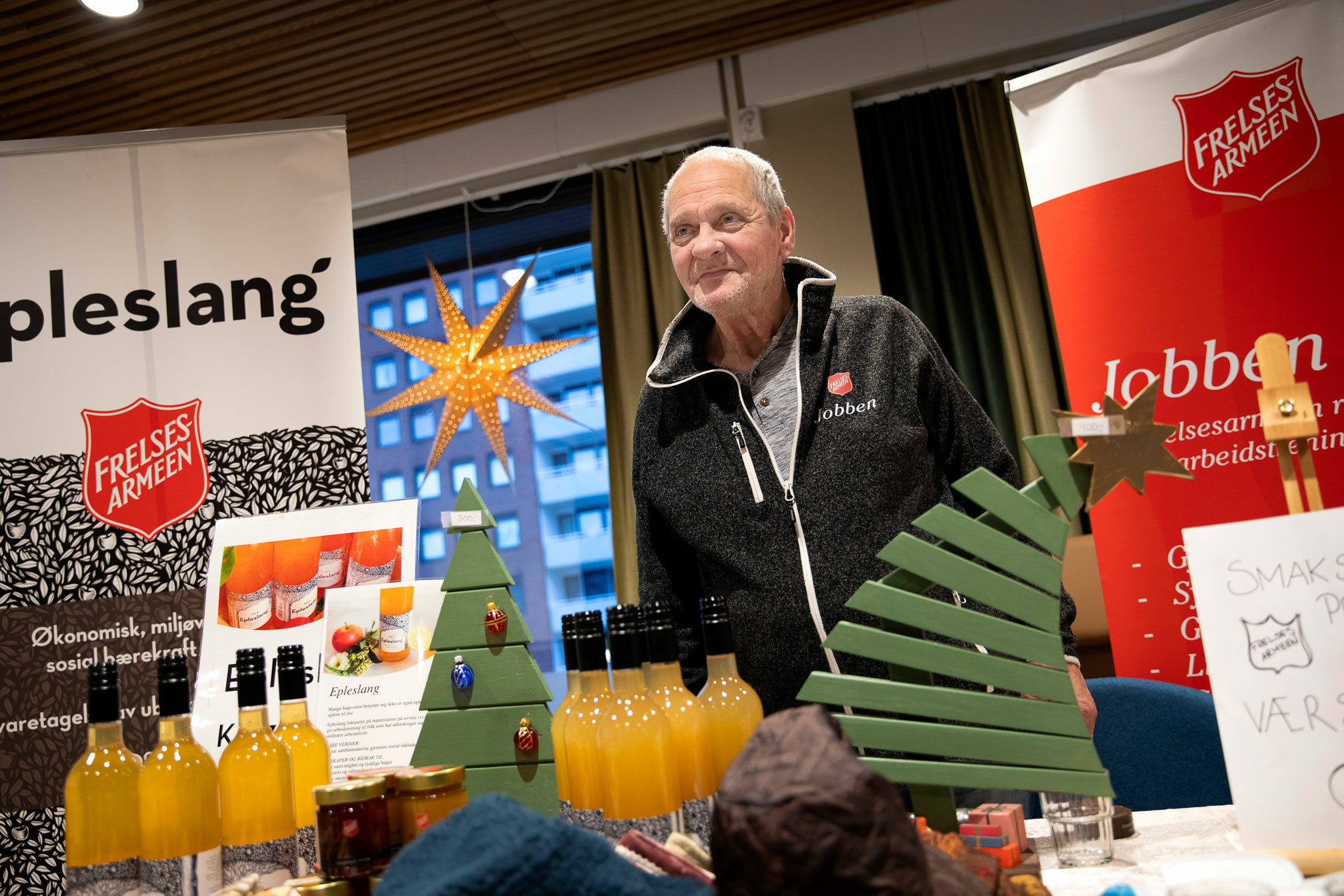 Foto: Eldre mann med logo fra Frelsesarmeen på jakka står bak  et bord og selger flasker og krukker. På bordet står også noen grønnmalte juletrær i tre.