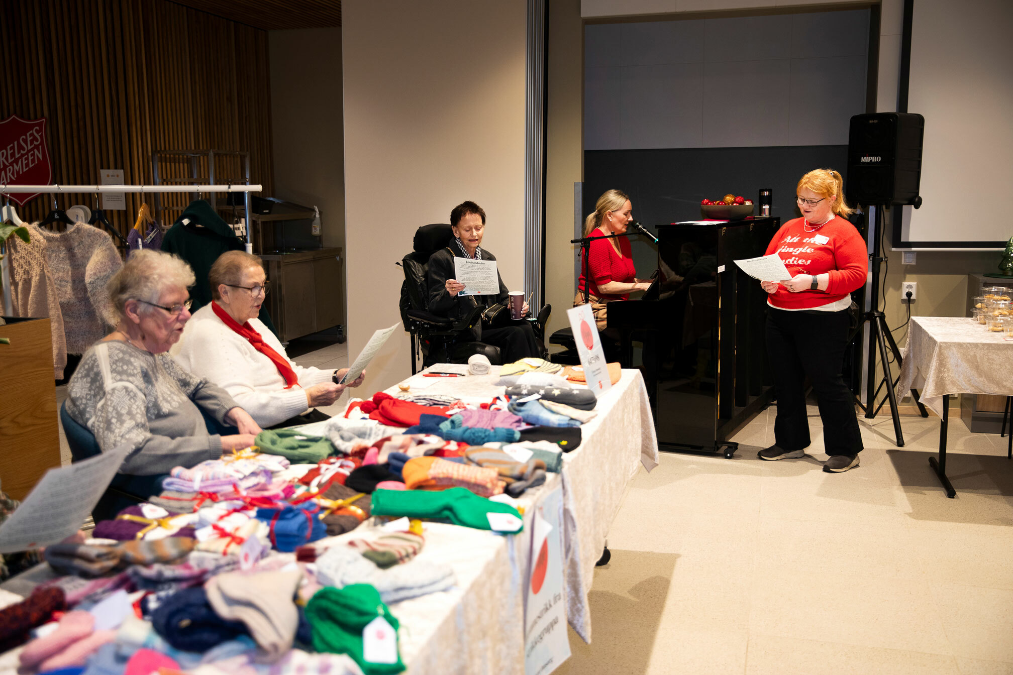 Foto: Det synges. En kvinne sitter ved pianoet og en annen står foran og synger. Rundt sitter eldre mennesker ved bord med hjemmestrikkede varer til salgs.