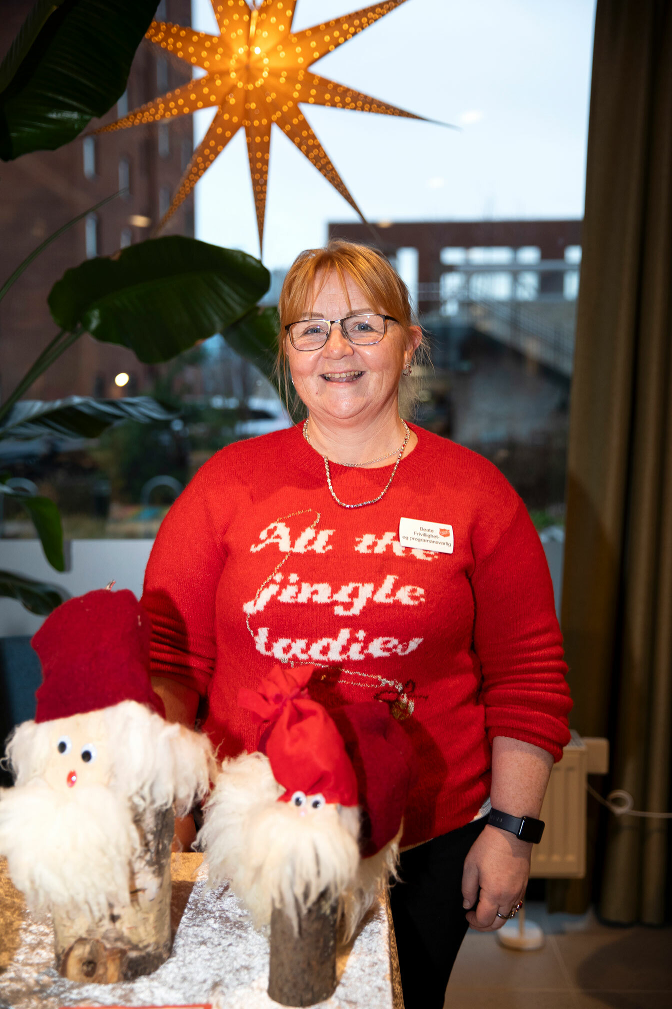 Foto: Kvinne i rød julegenser står bak noen nisser laget av trekubber med bark på. Bak henne henger en julestjerne.